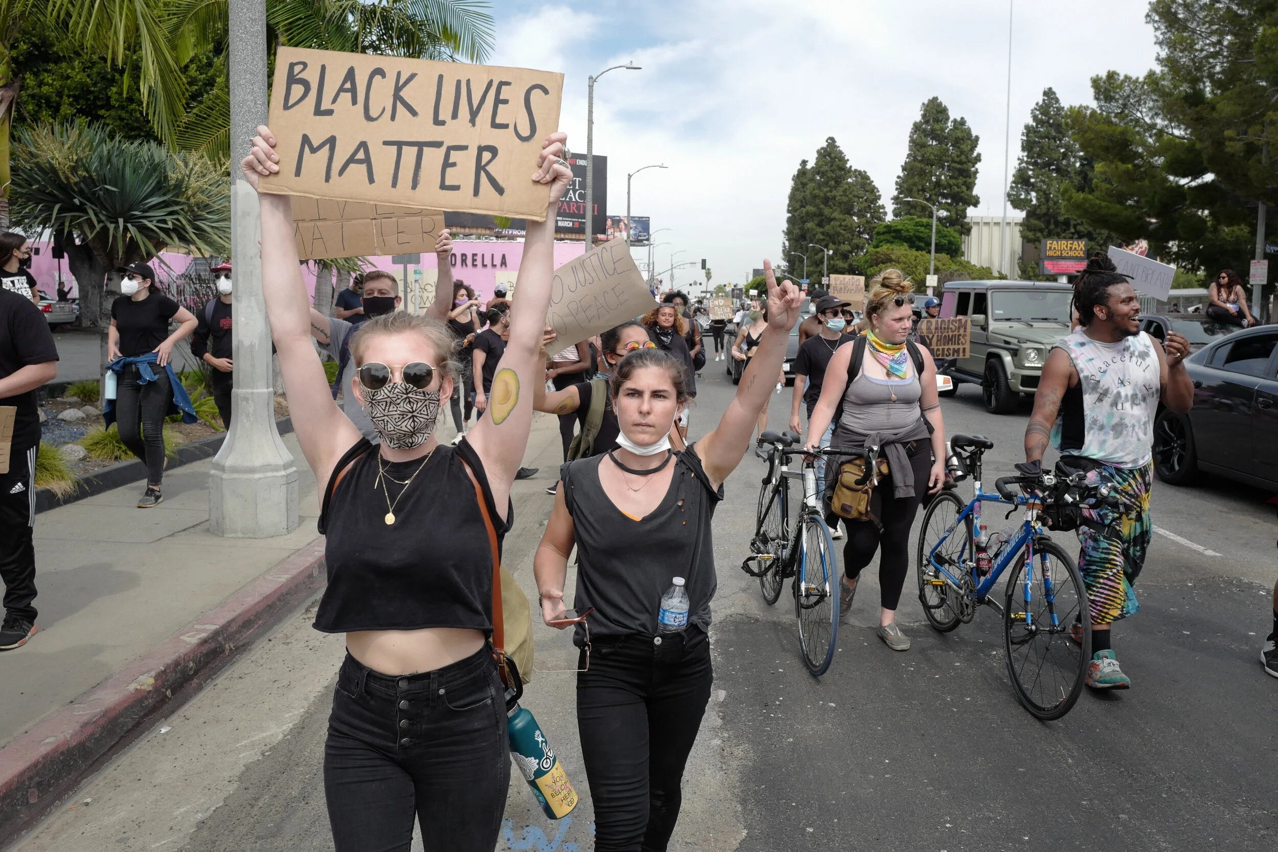 A group of protesters marching on a city street, with some holding signs, including one that reads 'Black Lives Matter' and others with messages for justice and peace. Some participants are on bicycles and wearing masks.