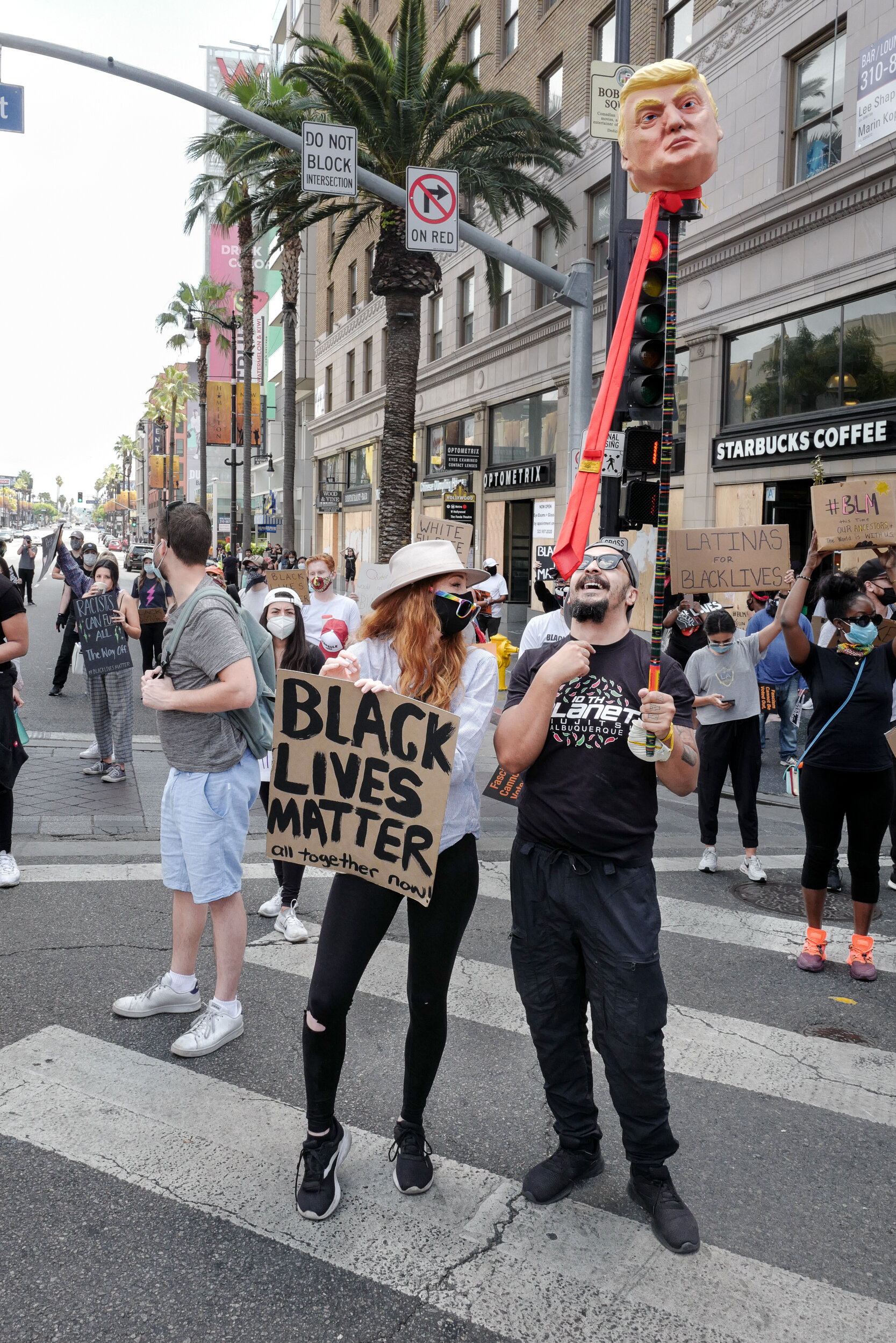 People participating in a protest march on a city street, holding signs supporting Black Lives Matter and other social justice causes, with a large inflatable figure of Donald Trump attached to a traffic light pole.