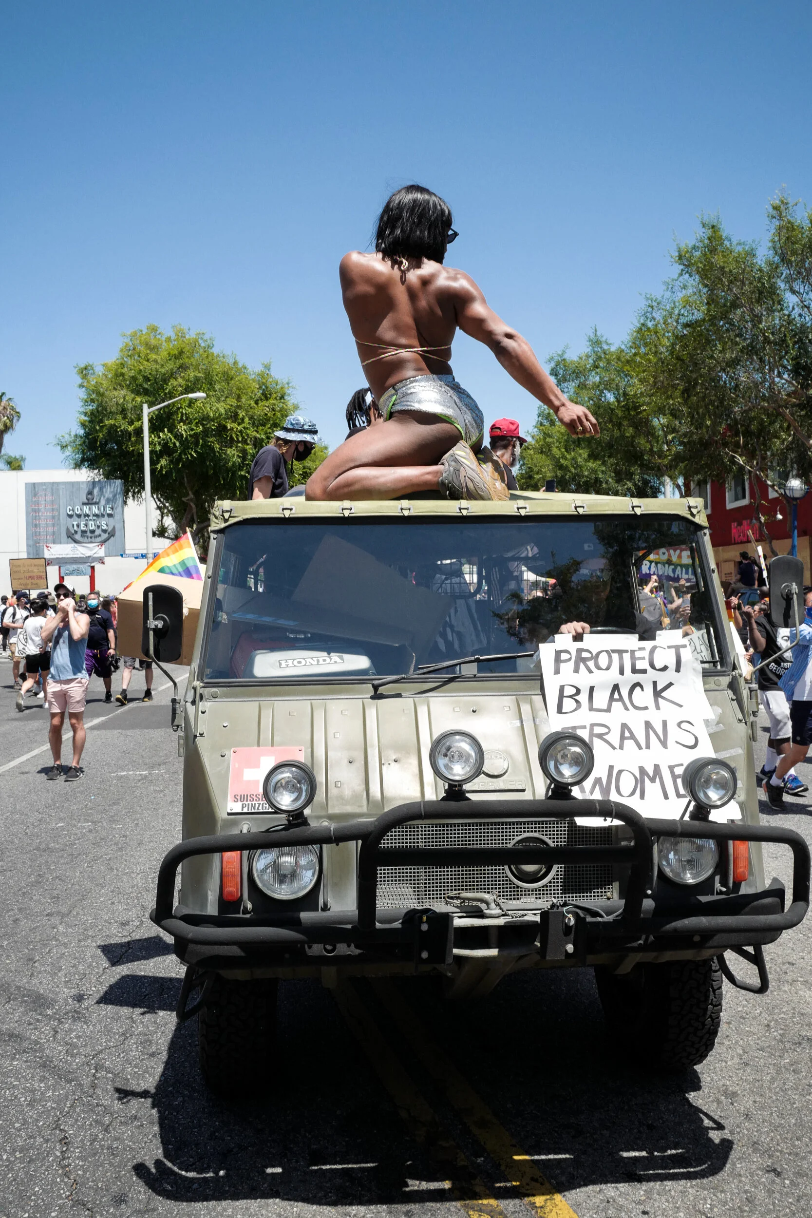 A person kneeling on top of a small military-style vehicle with a sign that reads "Protect Black Trans Women" during a protest or parade on a sunny street, with other protesters visible in the background.