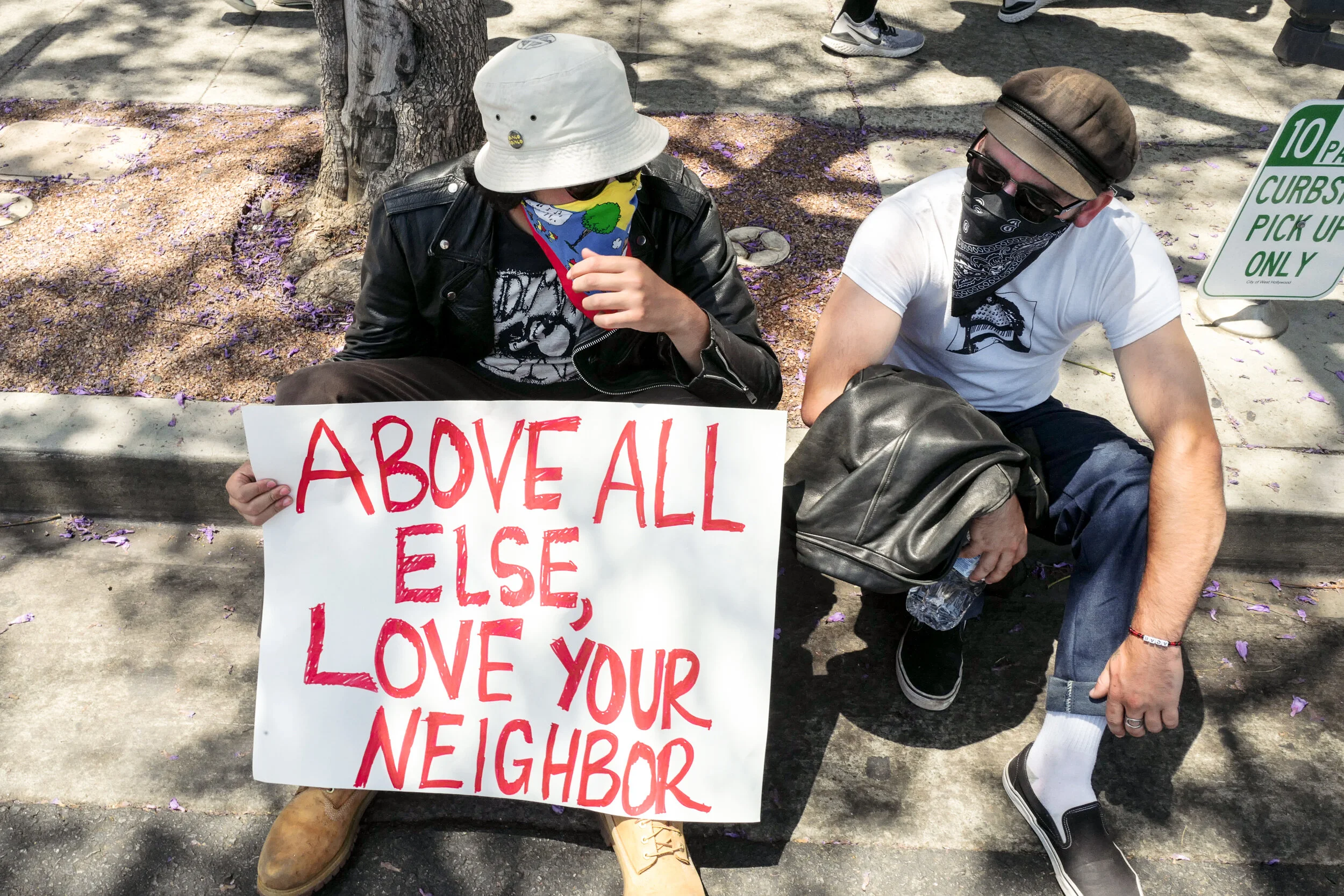 Two people sitting on the sidewalk, holding a sign that reads 'ABOVE ALL ELSE, LOVE YOUR NEIGHBOR'. They are wearing face coverings and casual clothing, with one person in a leather jacket and a white hat, and the other in a white t-shirt and a brown