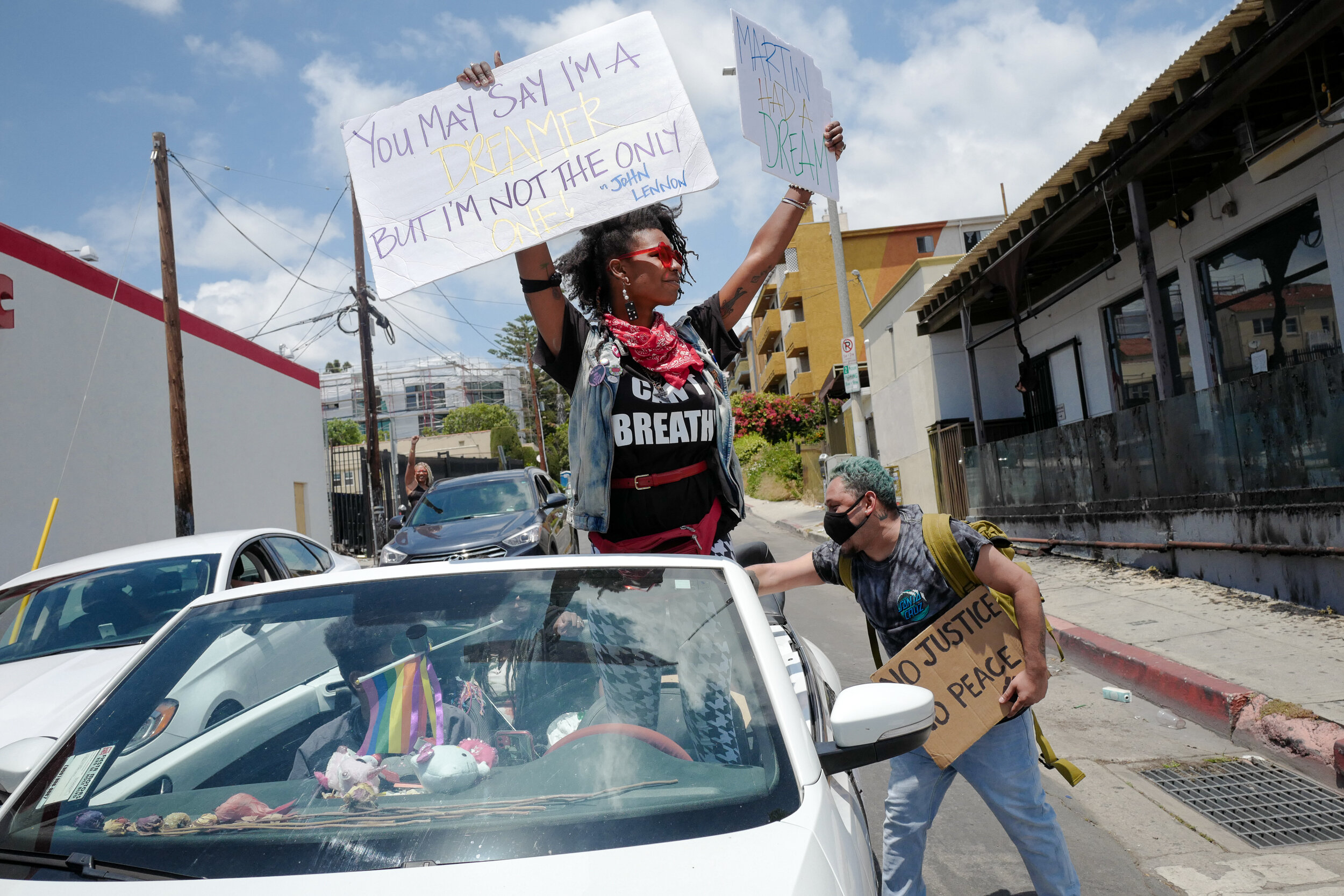 Woman standing in a white convertible car holding protest signs, one quoting John Lennon about dreaming, while a man with a backpack holds a sign that reads "No Justice No Peace". Other cars and buildings are in the background.