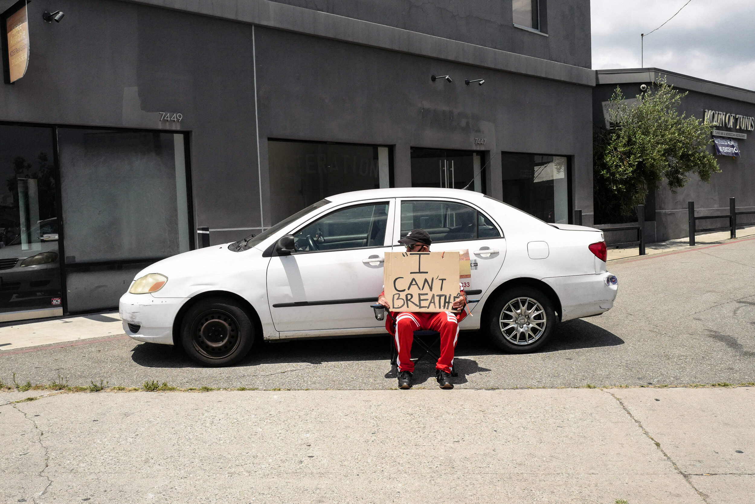 A person sitting on a folding chair in front of a white car on a city street, holding a cardboard sign that reads "I CAN'T BREATHE."