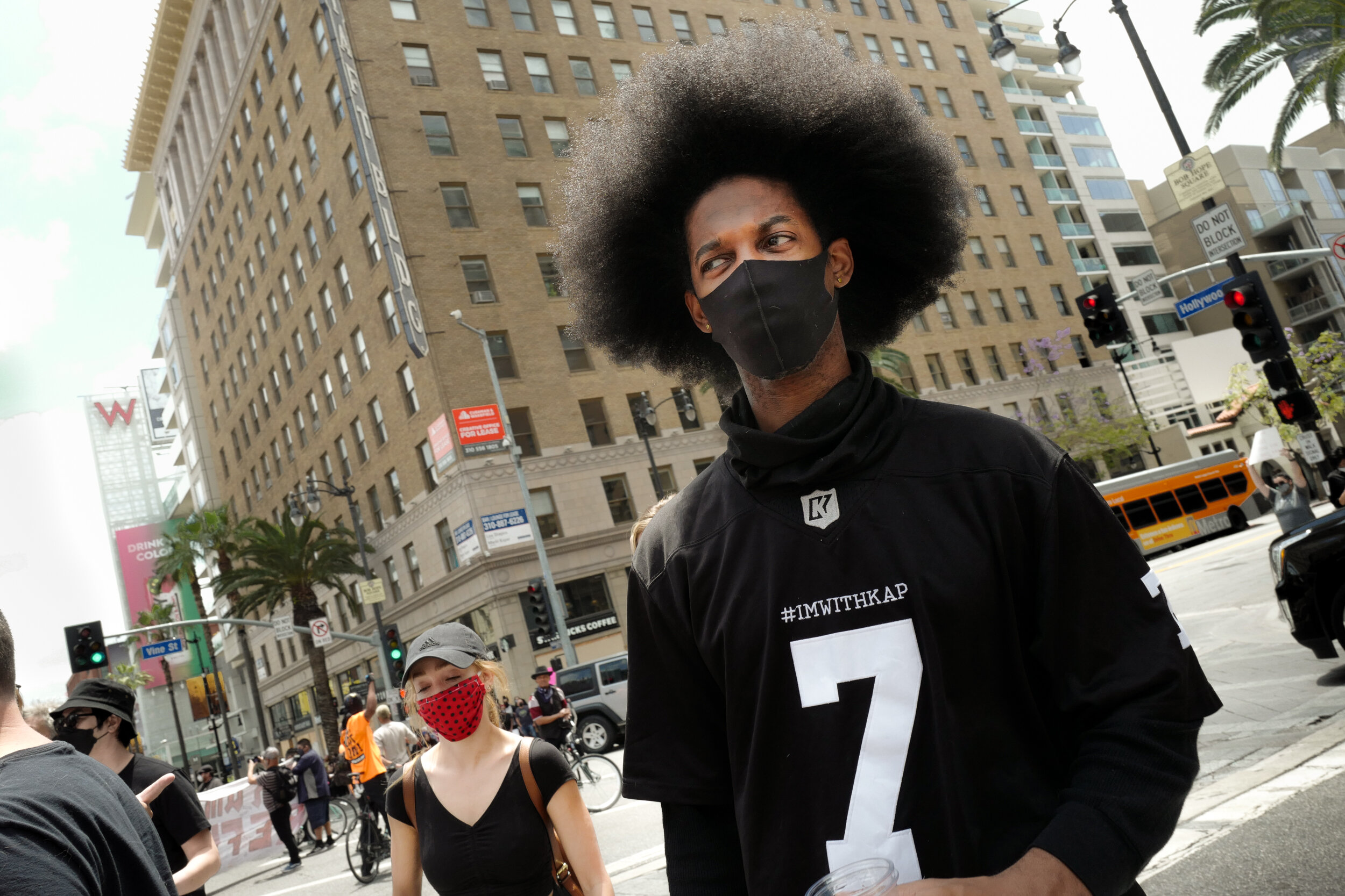 A young Black man with large afro hair wearing a black mask, black shirt with #IMWITHKAP and a large number 7, standing on a busy city street with people, buildings, traffic lights, and a yellow bus in the background.