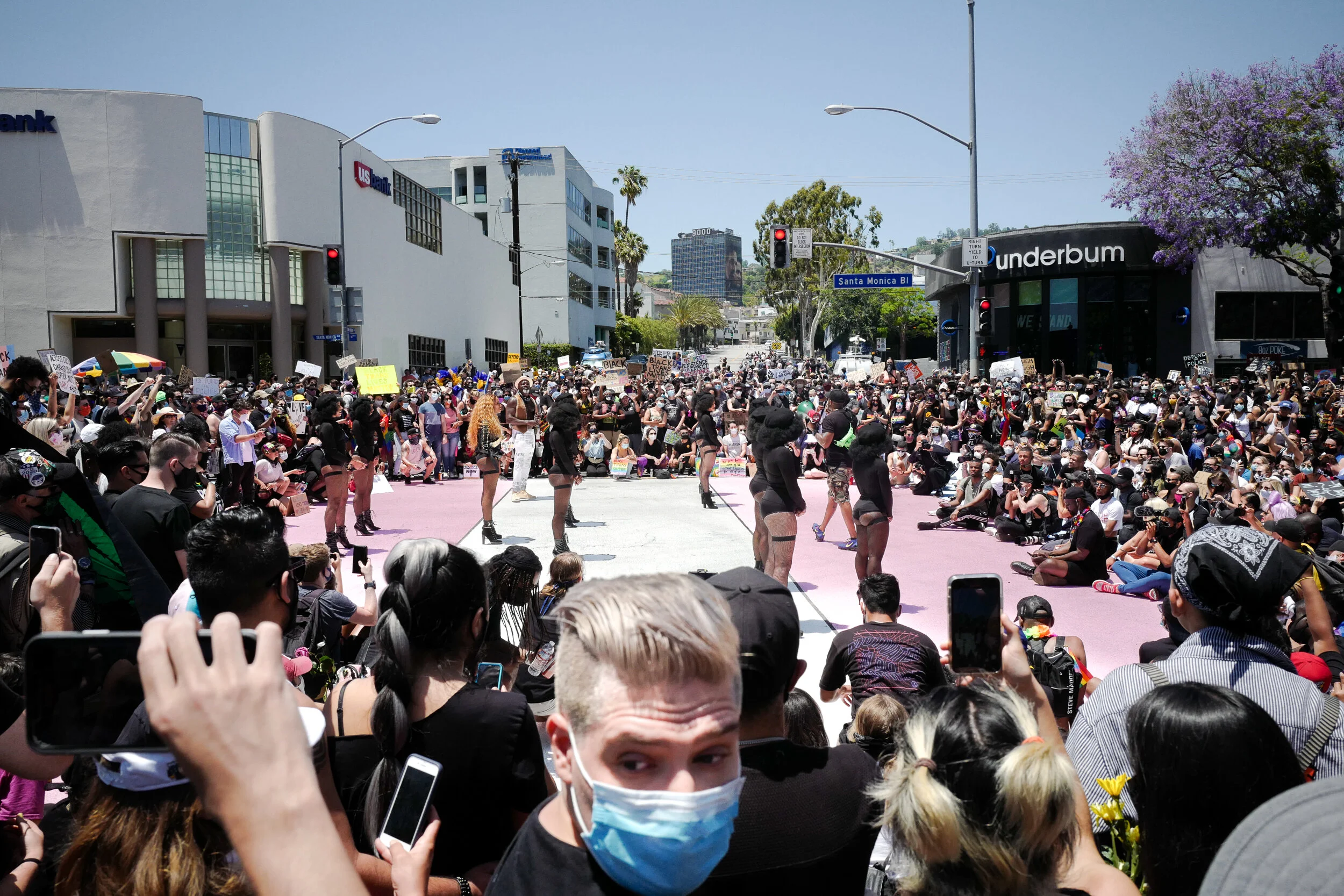 Crowd gathered at a protest or demonstration on a city street, with some people holding signs and taking photos, and a group of performers or dancers in the center.
