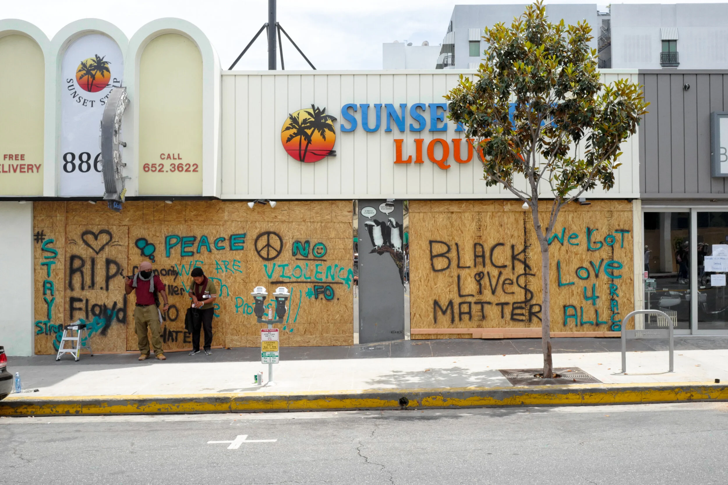 A storefront with a 'Sunset Liquor' sign, boarded-up windows with graffiti including peace, love, and Black Lives Matter messages, two people standing on the sidewalk, and a tree in front of the building.