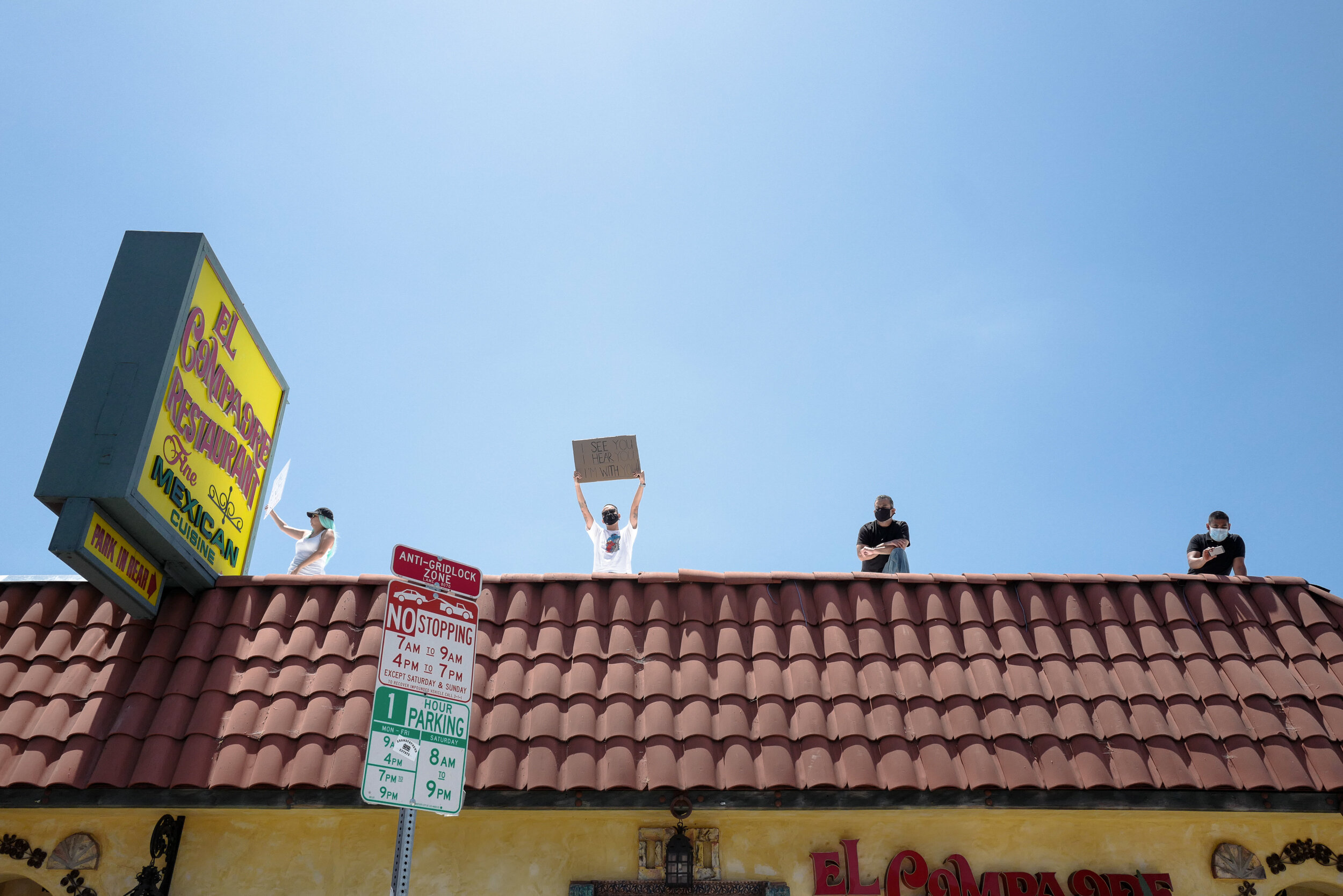 Four people wearing masks on a restaurant roof, two holding signs, under a clear blue sky.