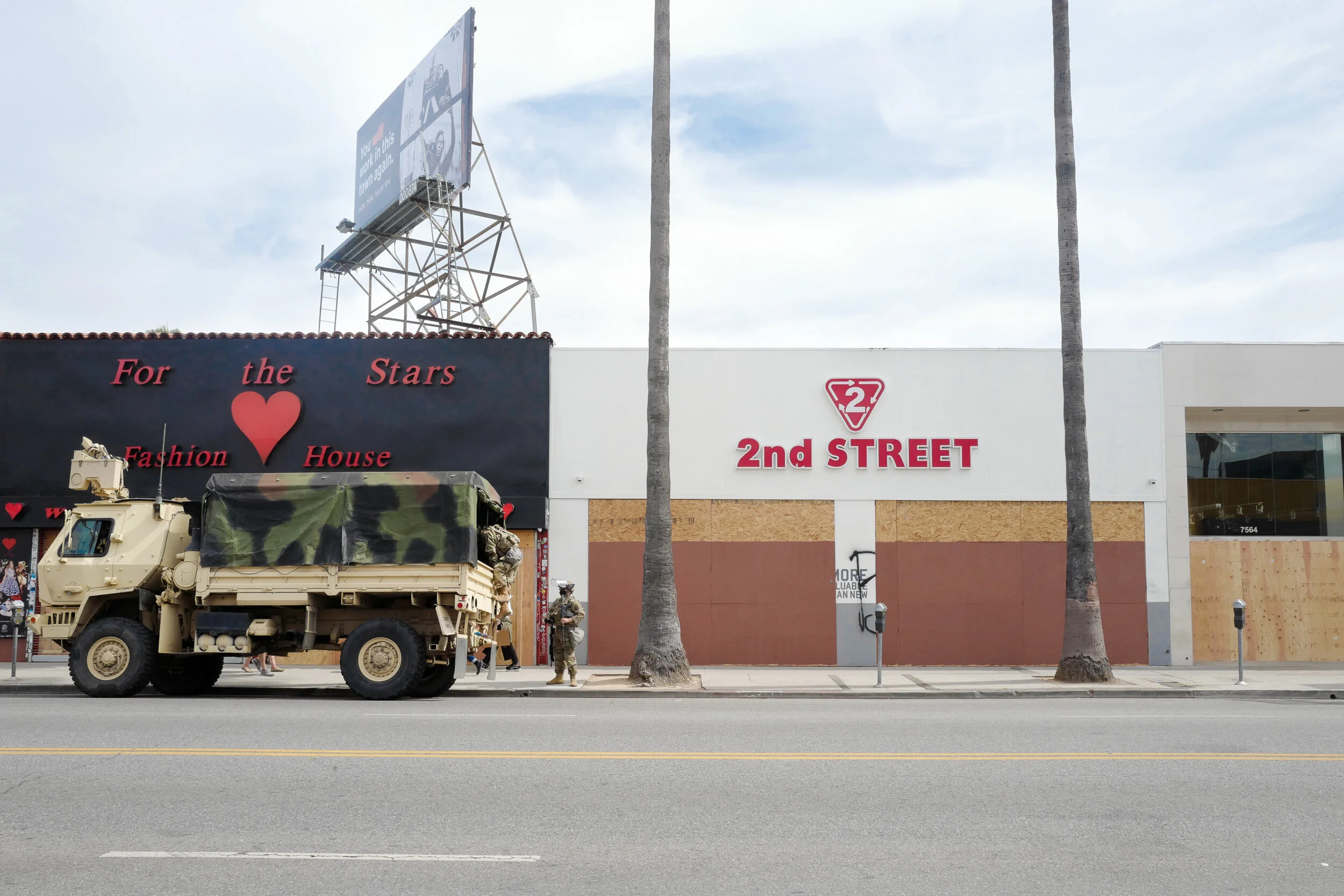 A beige military vehicle parked on the street in front of a building with signs including 'For the Stars,' a red heart, and '2nd STREET.' There are soldiers and pedestrians nearby, and palm trees along the sidewalk.