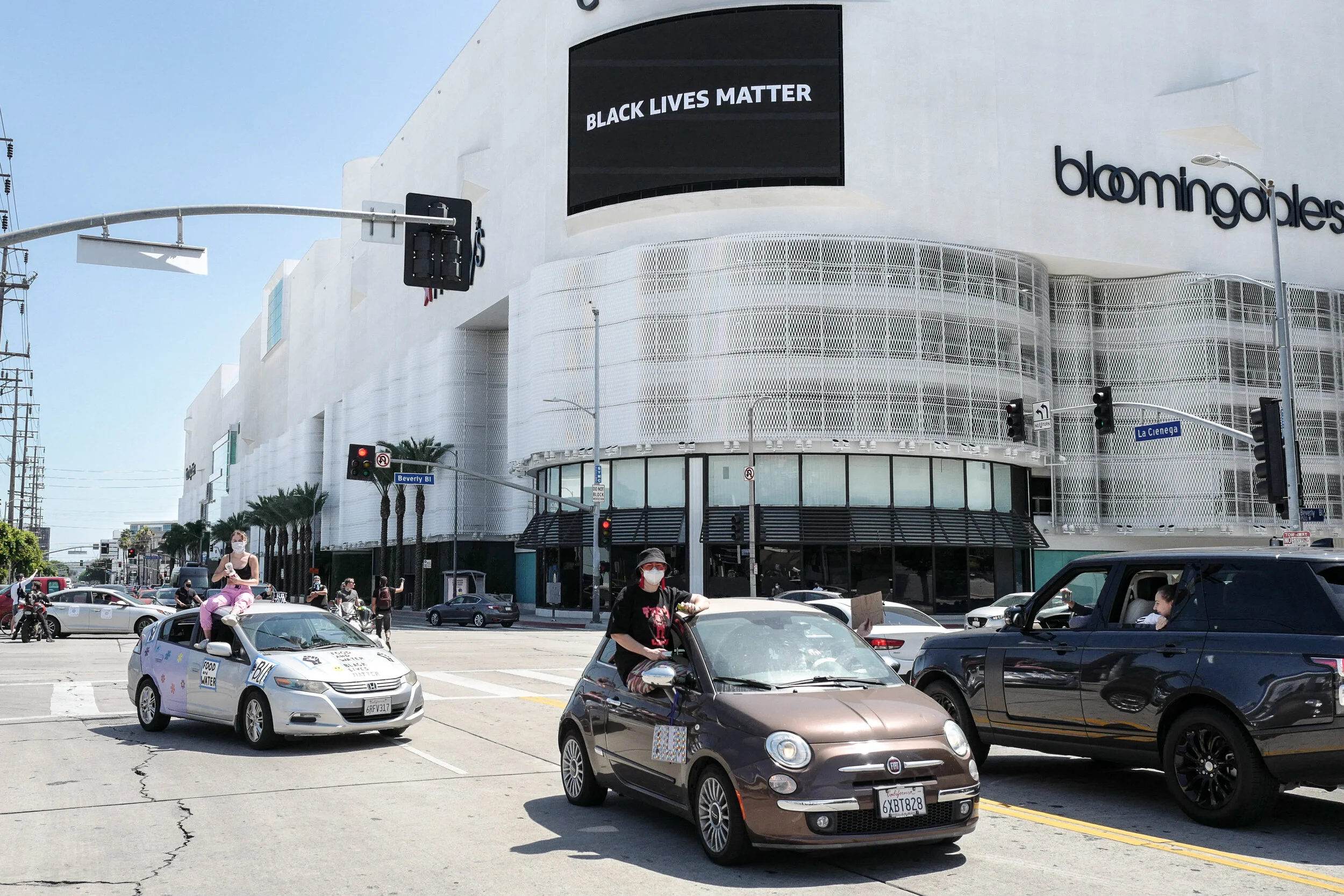 Street scene in Los Angeles with people in cars, some with people sitting on rooftops, and a large white building with a sign saying 'Black Lives Matter' and the Bloomingdale's store.