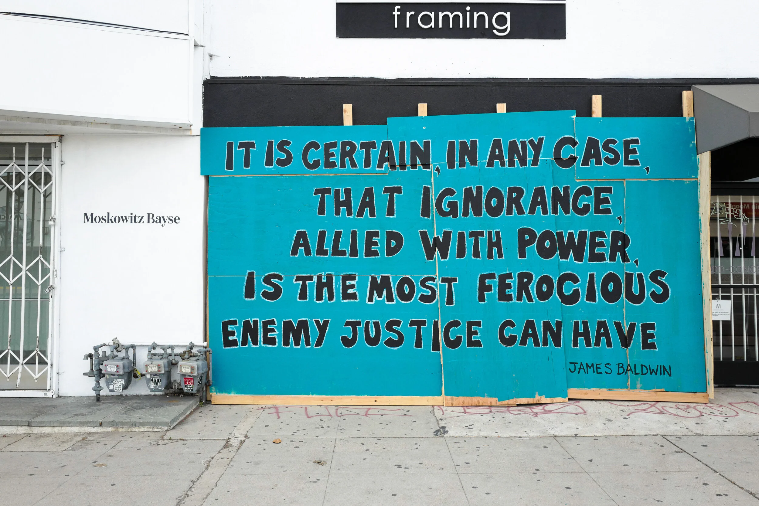 Large blue protest sign with a quote by James Baldwin that says, 'It is certain, in any case, that ignorance allied with power is the most ferocious enemy justice can have.' The sign is placed on a sidewalk, with a sign that says 'Moskowitz Bayse' to