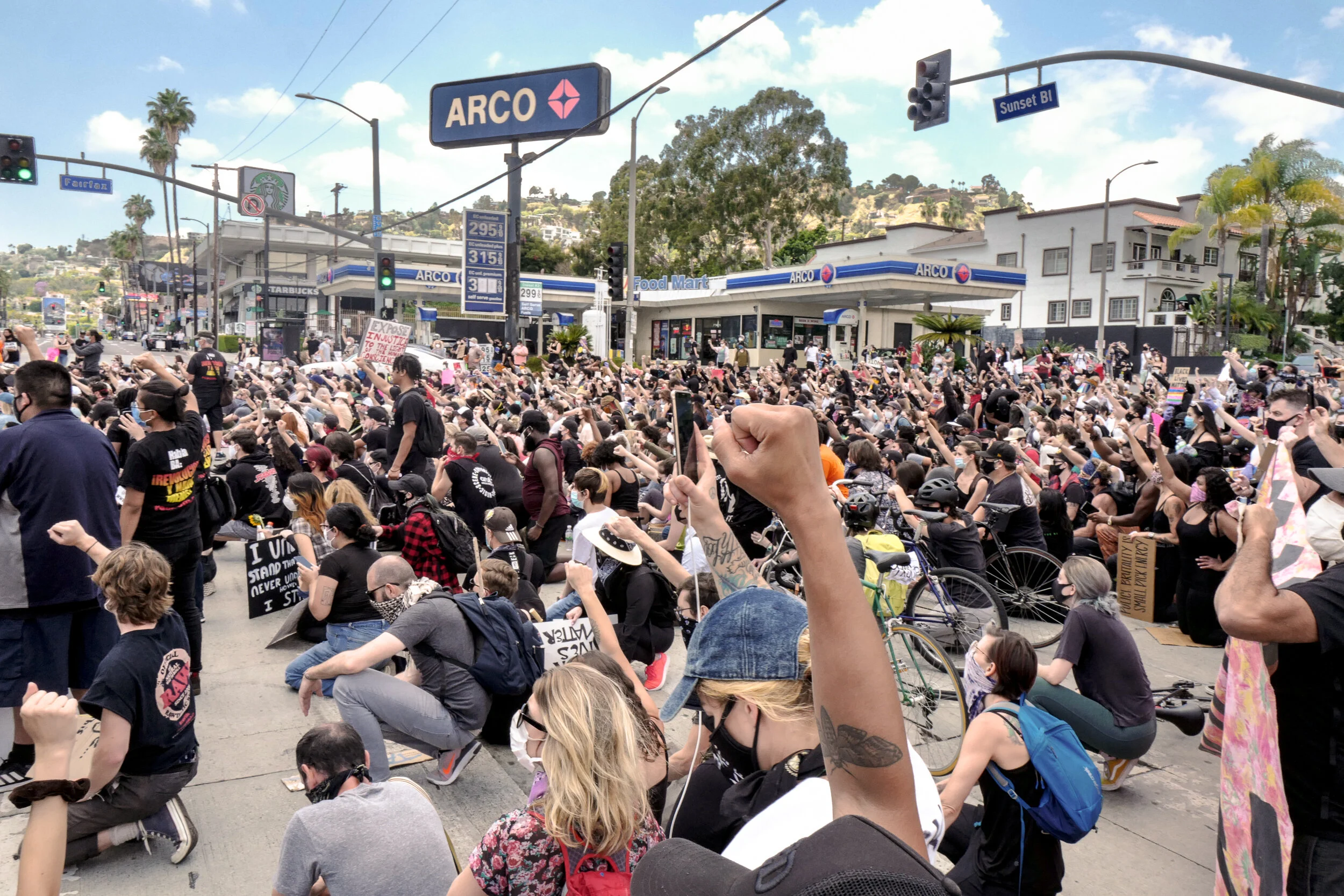 Large crowd of protesters sitting and kneeling on the street, many with fists raised, in front of an ARCO gas station and Starbucks. Some people are holding signs, and many are wearing masks.