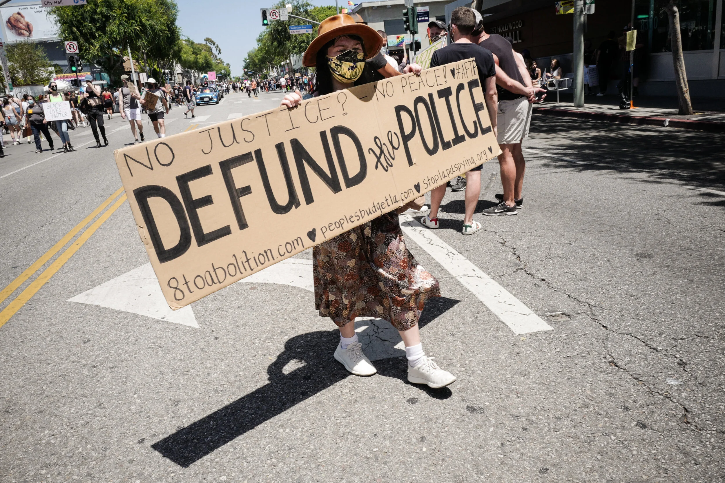 Protestor holding a large cardboard sign at a street protest. The sign reads 'No Justice? No Peace! Defund the Police' with websites and message details. Other protesters and signs are visible in the background.