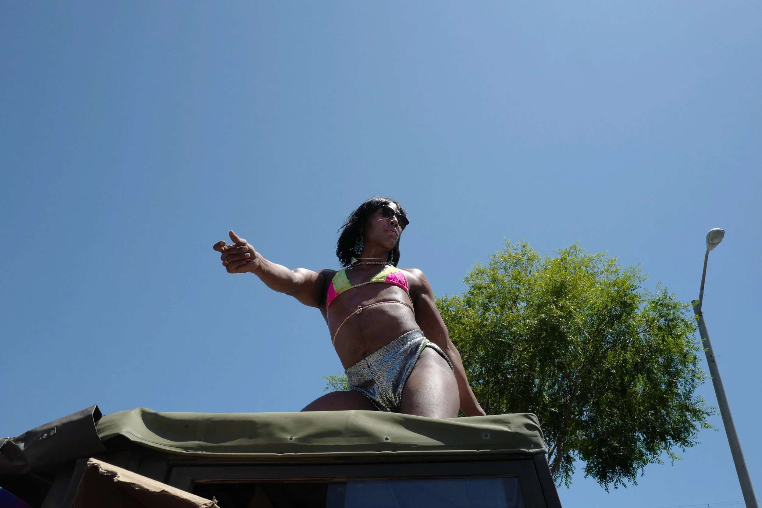 A person with dark hair, sunglasses, and decorated in colorful clothing is standing on top of a vehicle, pointing with one hand, against a clear blue sky and green trees in the background.