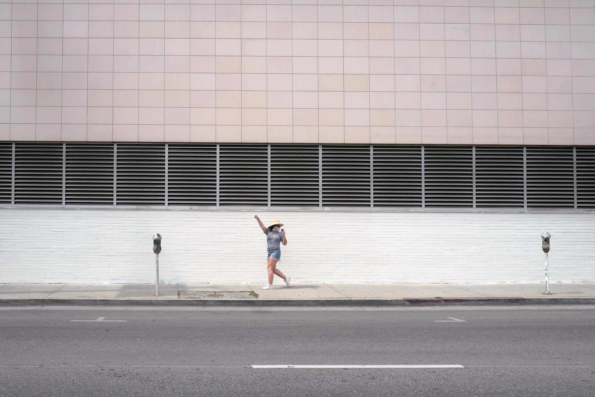 A person walking on the sidewalk in front of a building wall, wearing a hat, face mask, grey t-shirt, shorts, and sneakers, holding a phone, with parking meters visible.