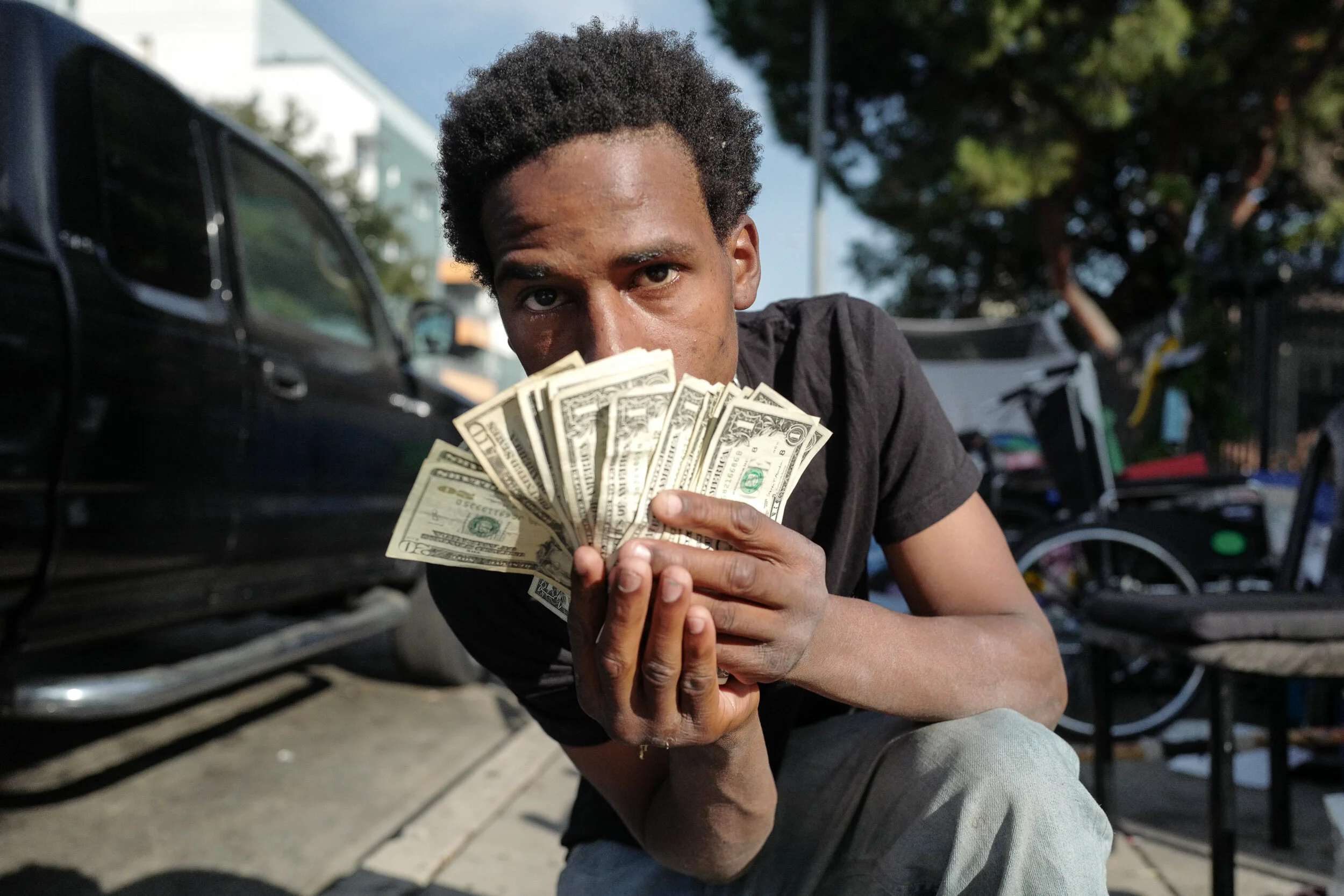 A young man kneels on the sidewalk holding and displaying a fanned-out stack of U.S. dollar bills, looking directly at the camera with a serious expression, with parked cars and trees in the background.