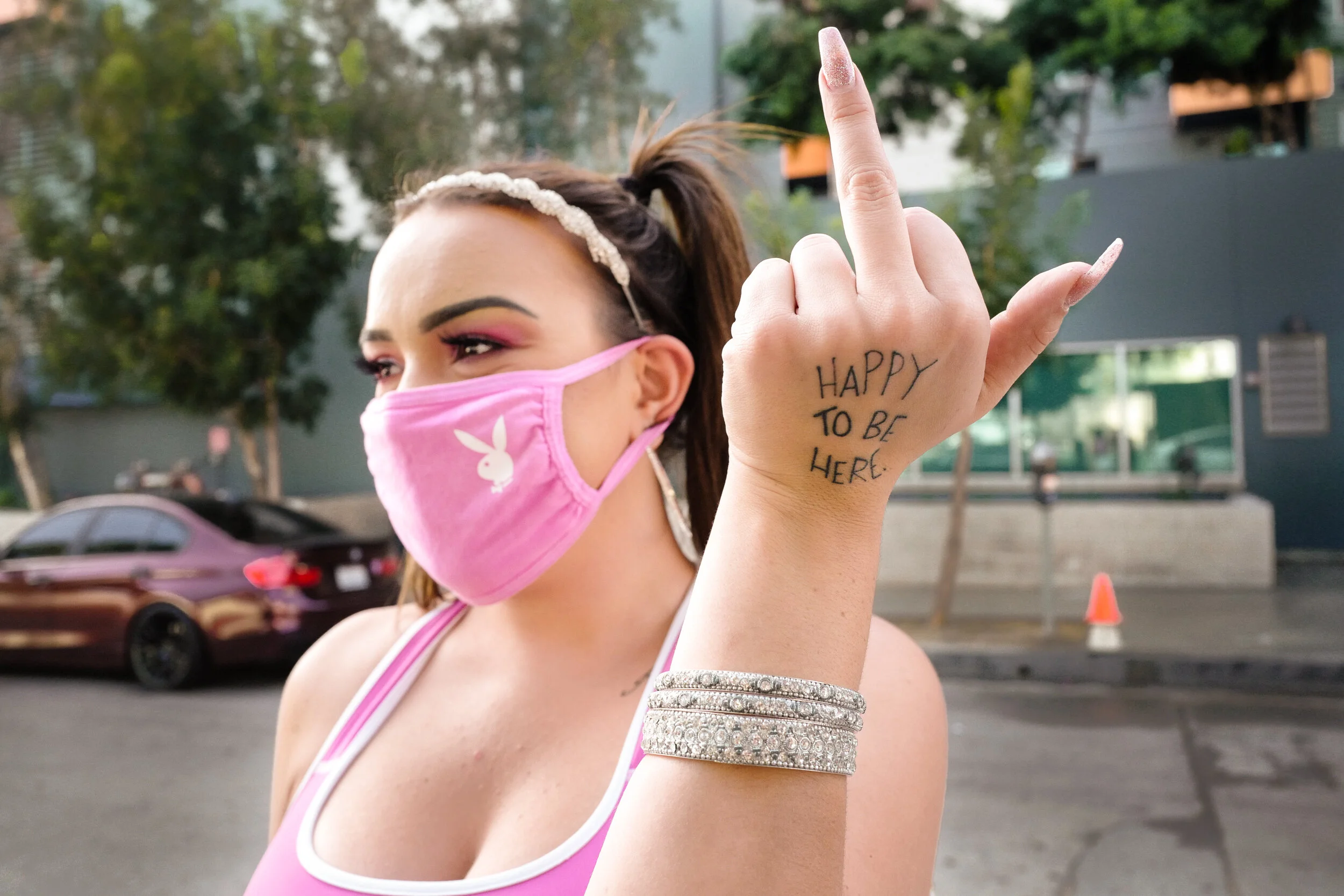 A woman wearing a pink face mask with a Playboy bunny logo, earrings, and a rhinestone bracelet, showing her defiantly making a hand gesture with the words "HAPPY TO BE HERE" written on her hand, standing outdoors on a city street.