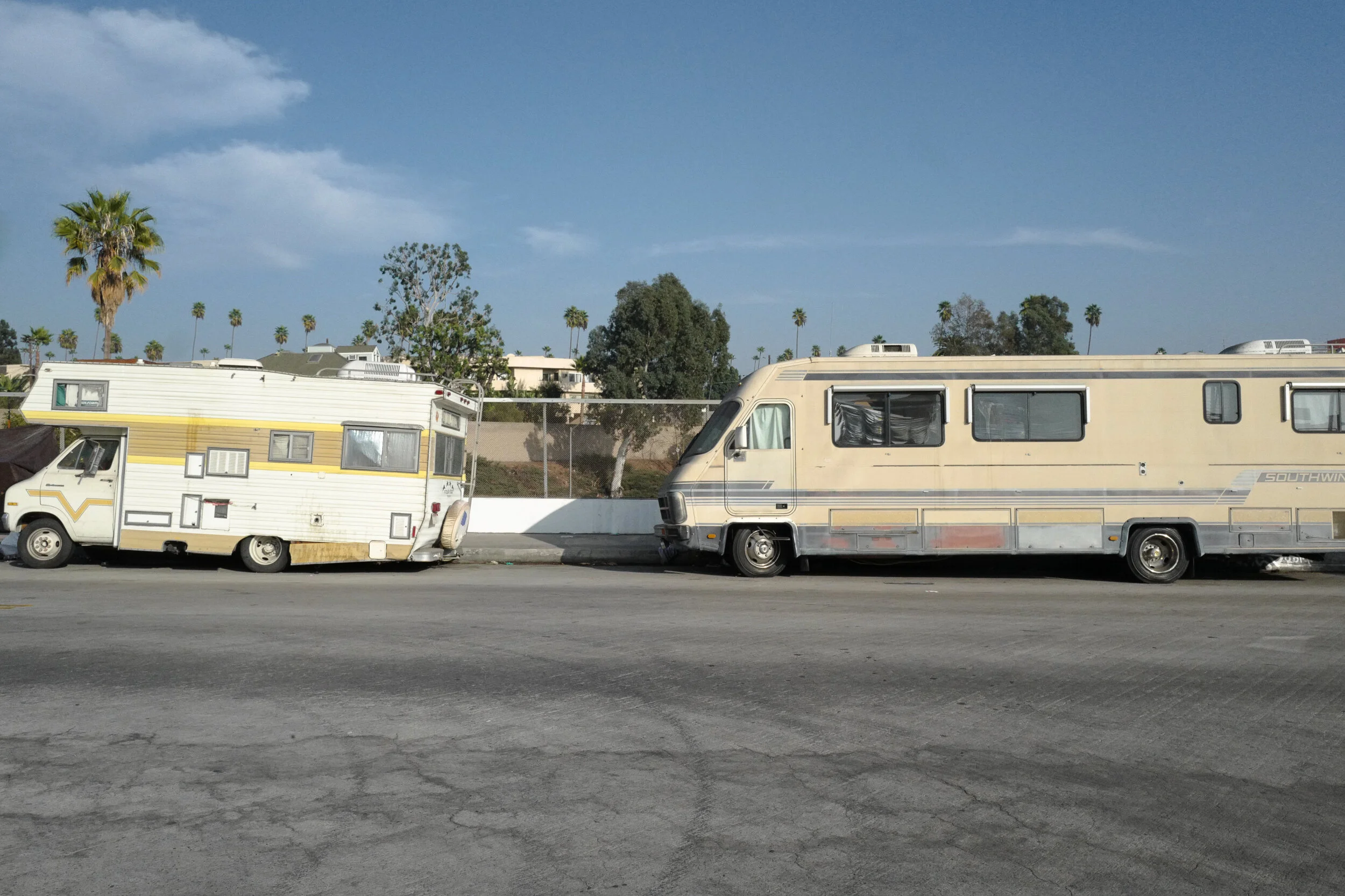 Two old recreational vehicles parked on a street with palm trees and residential houses in the background. The sky is partly cloudy.