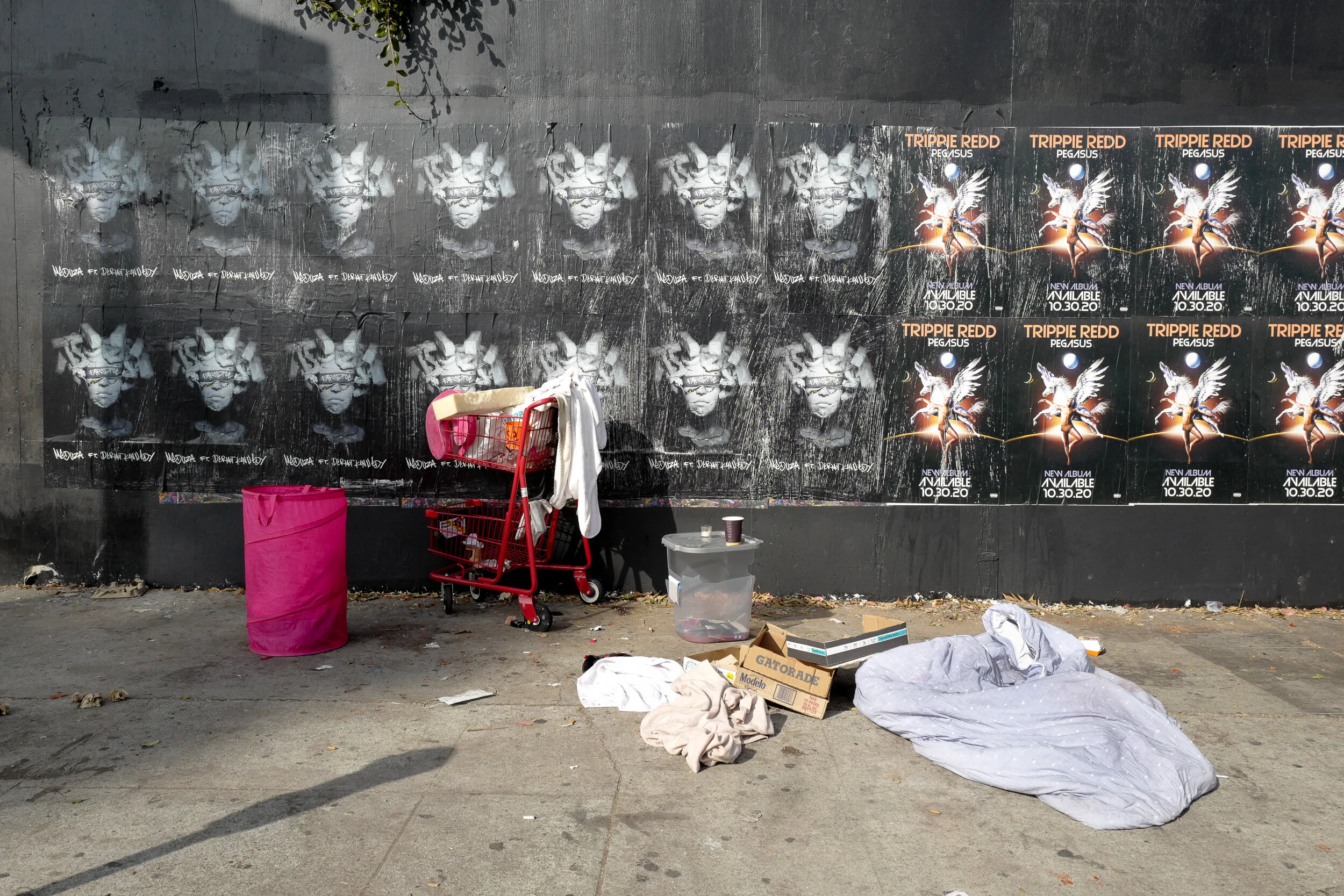 Street scene with trash and posters on the wall, including artwork of a face wearing glasses and advertisements for a Trippie Redd album.