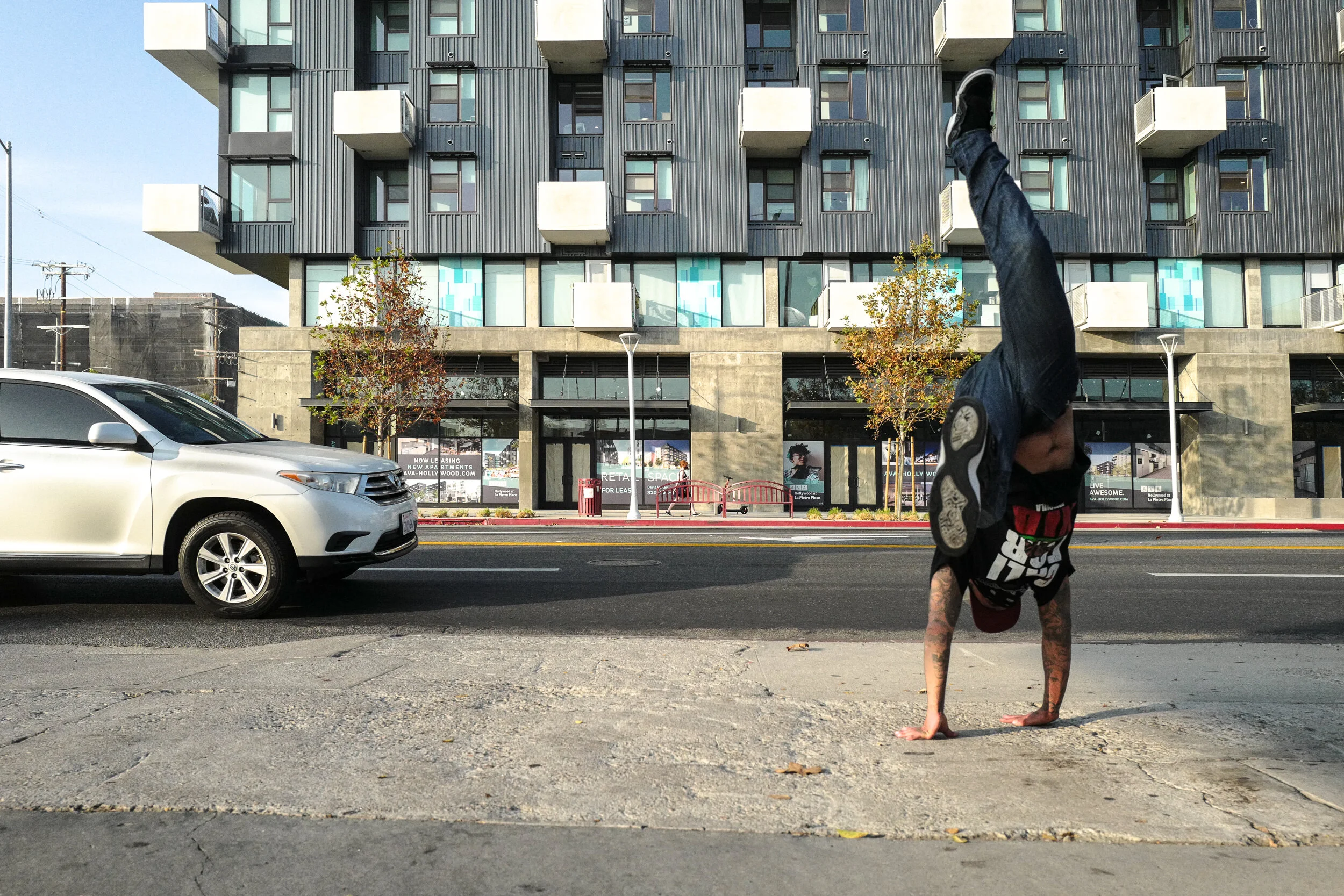 A person performing a handstand on the sidewalk with a city street and modern apartment building in the background.