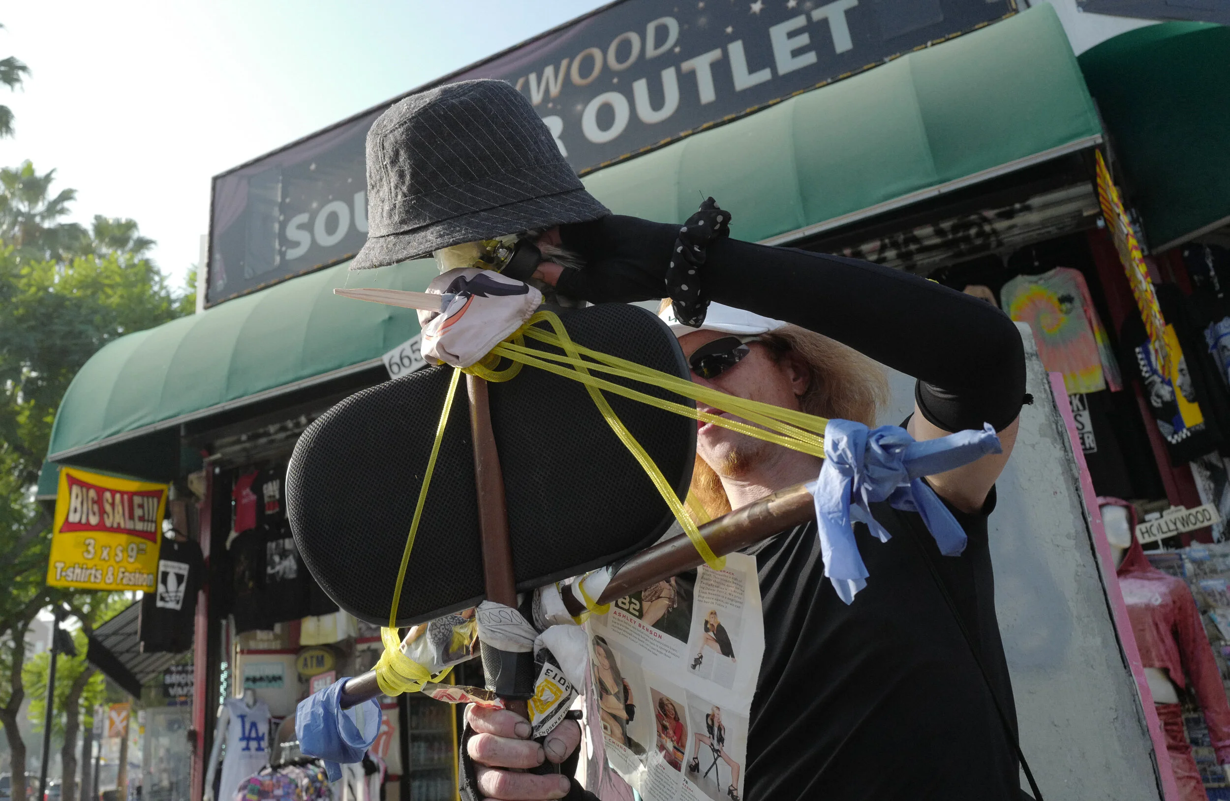 A person with long red hair, wearing dark sunglasses, is decorating a mannequin with a black hat, black gloves, and a newspaper, outside a souvenir shop with a green awning and sale signs.