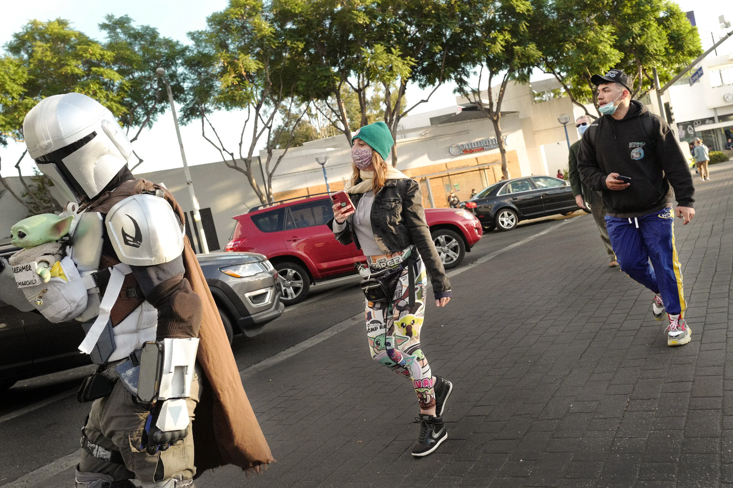 Person dressed as a Star Wars character talking to a woman and a man walking on the sidewalk. The woman is using her phone, and all three are wearing masks.