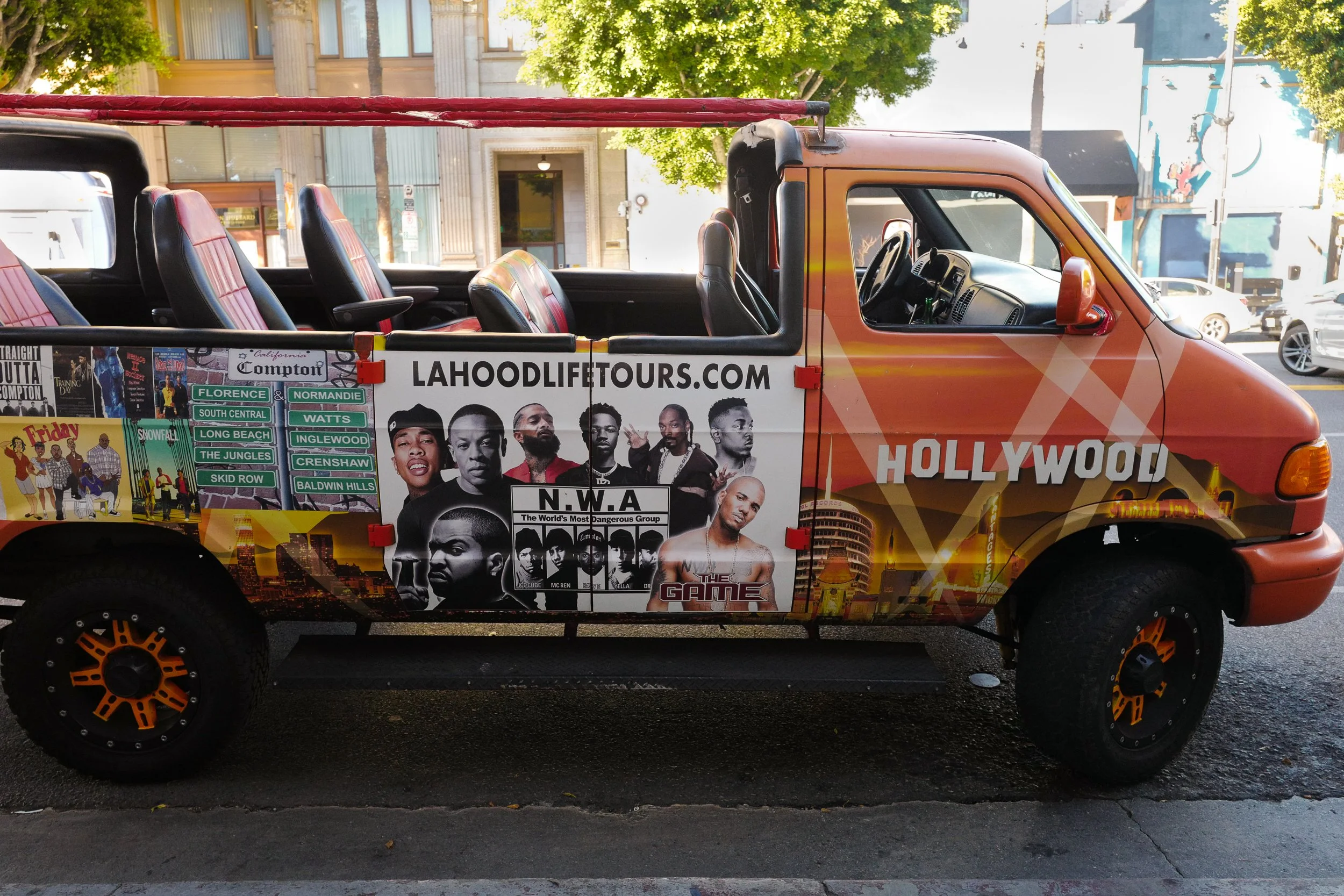 A colorful vehicle decorated with various posters and photographs, including images of hip-hop artists, cityscapes, and website information, parked on a city street with trees and buildings in the background.