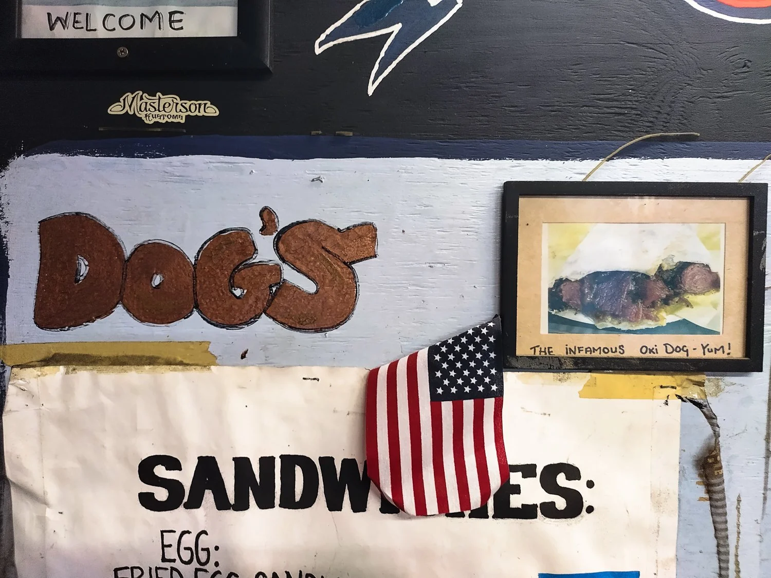 Decorative wall with a sign that says "DOGS" in large brown lettering, a framed picture of a grilled hot dog with mustard labeled "The Infamous Oki Dog-Yum!", a small American flag patch, and a partially visible menu with the word "SANDWICHES".