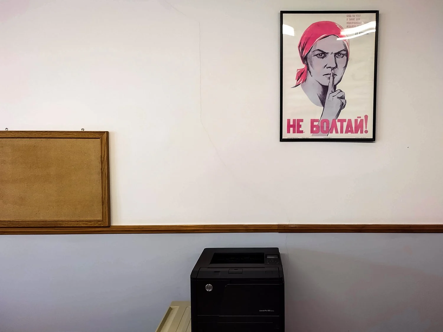 A framed poster on a white wall featuring a woman with a red headscarf and finger to lips, with Russian text that reads "Не болтай!" (“Don’t chatter!”).