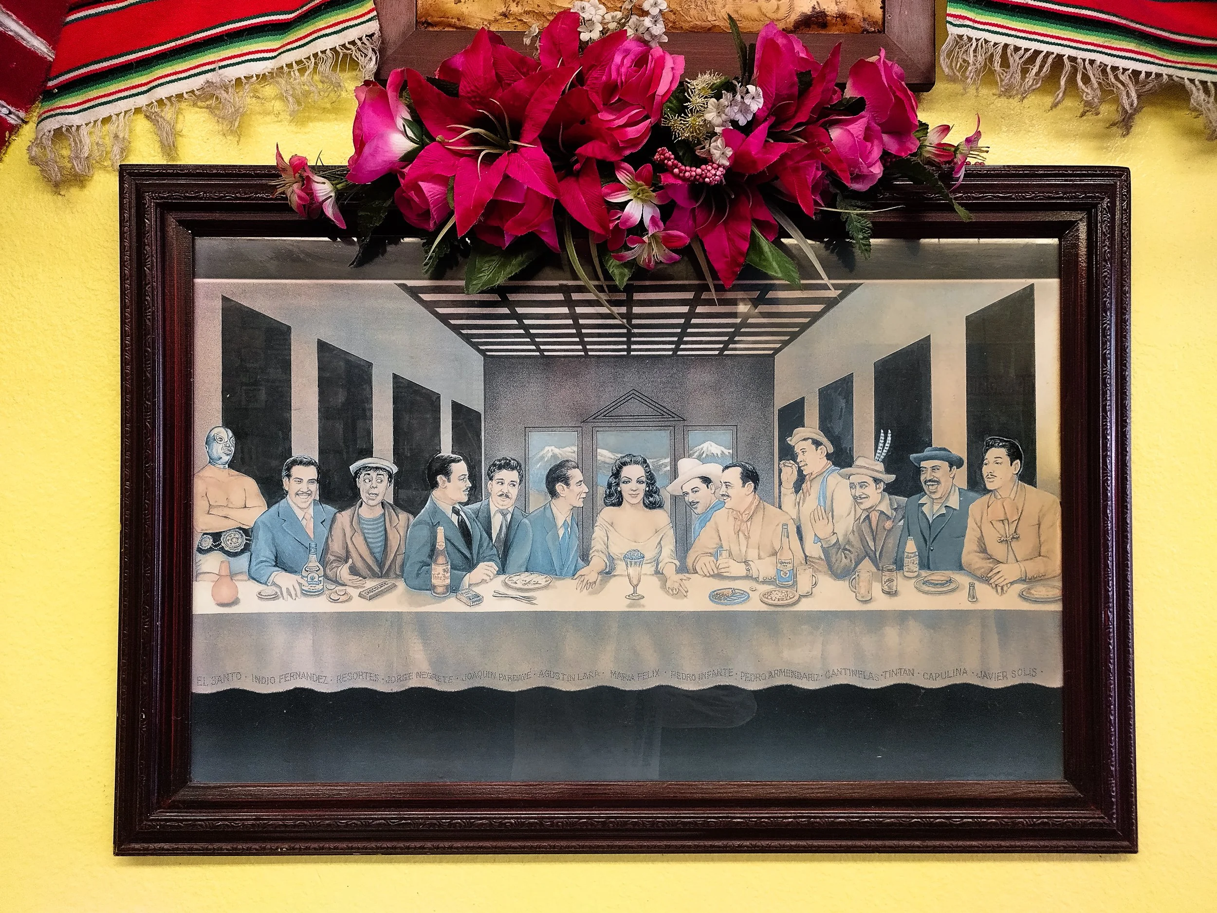A framed picture of the famous painting "El Mural de la Última Cena" with a flower arrangement on top, hanging on a yellow wall. The painting depicts a group of people sitting at a dining table, some talking, some drinking, with a mountainous landsca