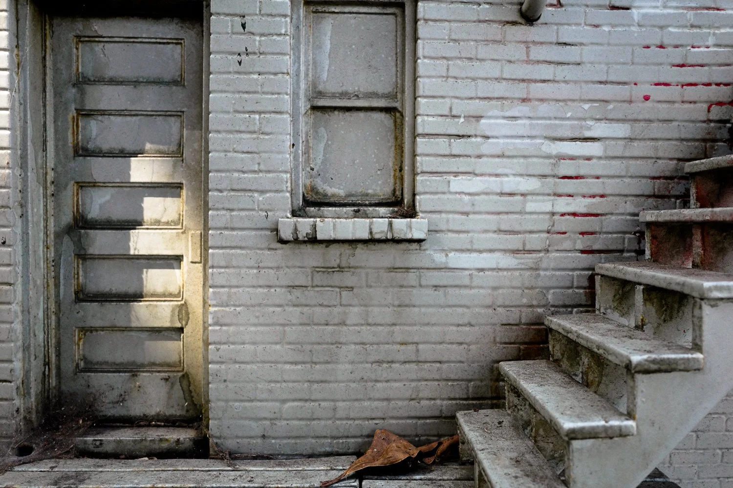 A weathered, white brick wall with a small window and a rusty staircase on the right, next to an old metal door with a vertical row of rectangular glass panels, and a dried leaf on the ground.