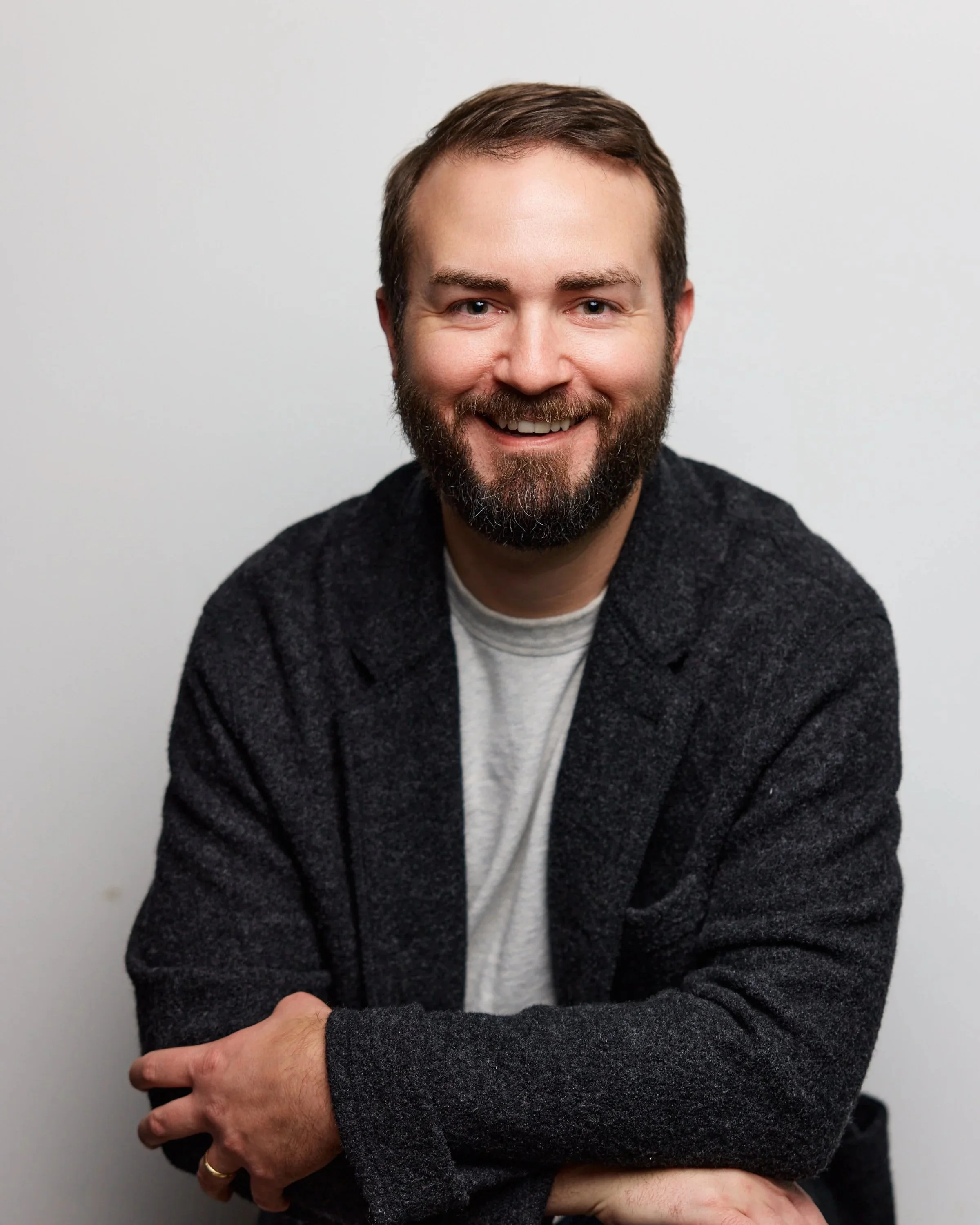 A man with a beard and short brown hair, smiling, wearing a dark sweater over a light shirt, is sitting with his arms crossed against a plain white background.