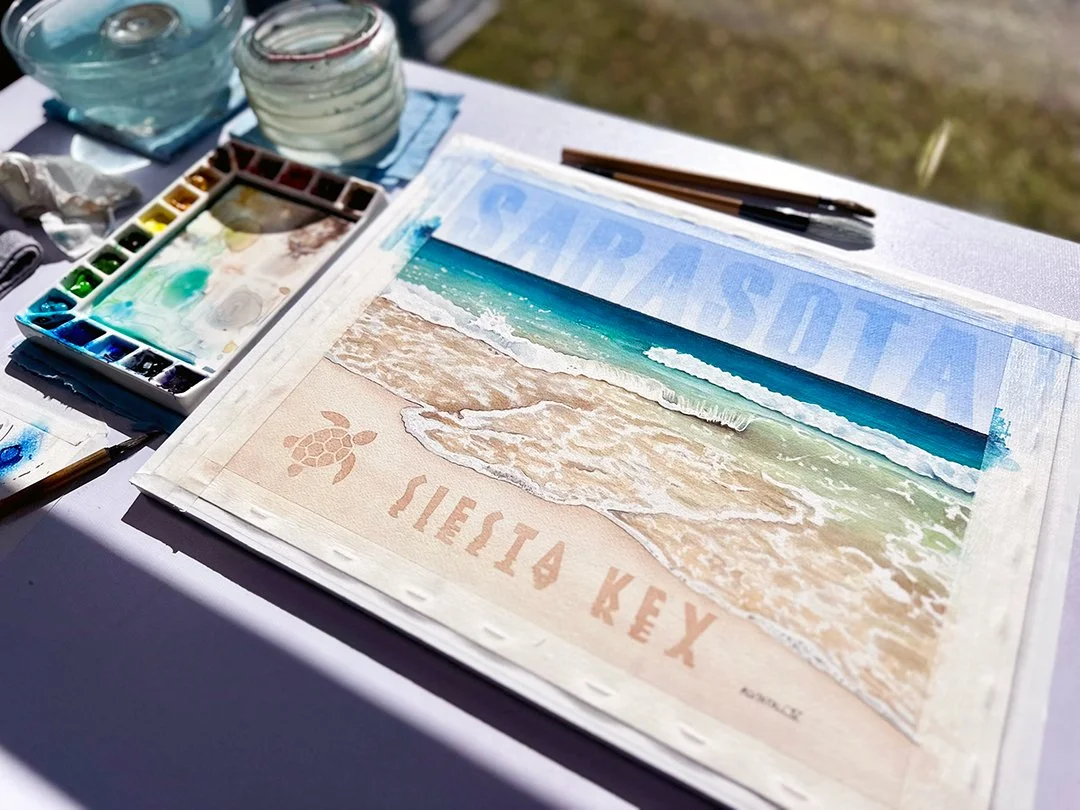 Finished Siesta Key Beach watercolor painting on artist's desk with watercolor palette, water jars, and brushes showing the creative process.
