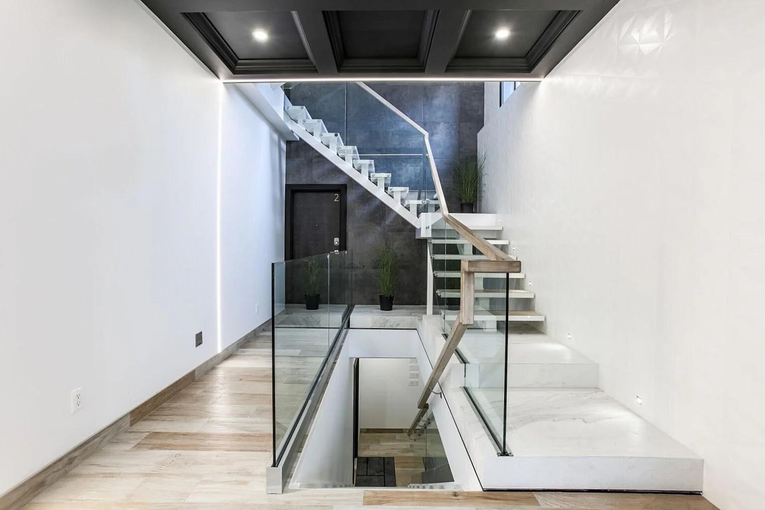 Modern staircase with glass railing and potted plants at the top, leading to a lower level in a contemporary, minimalist interior.