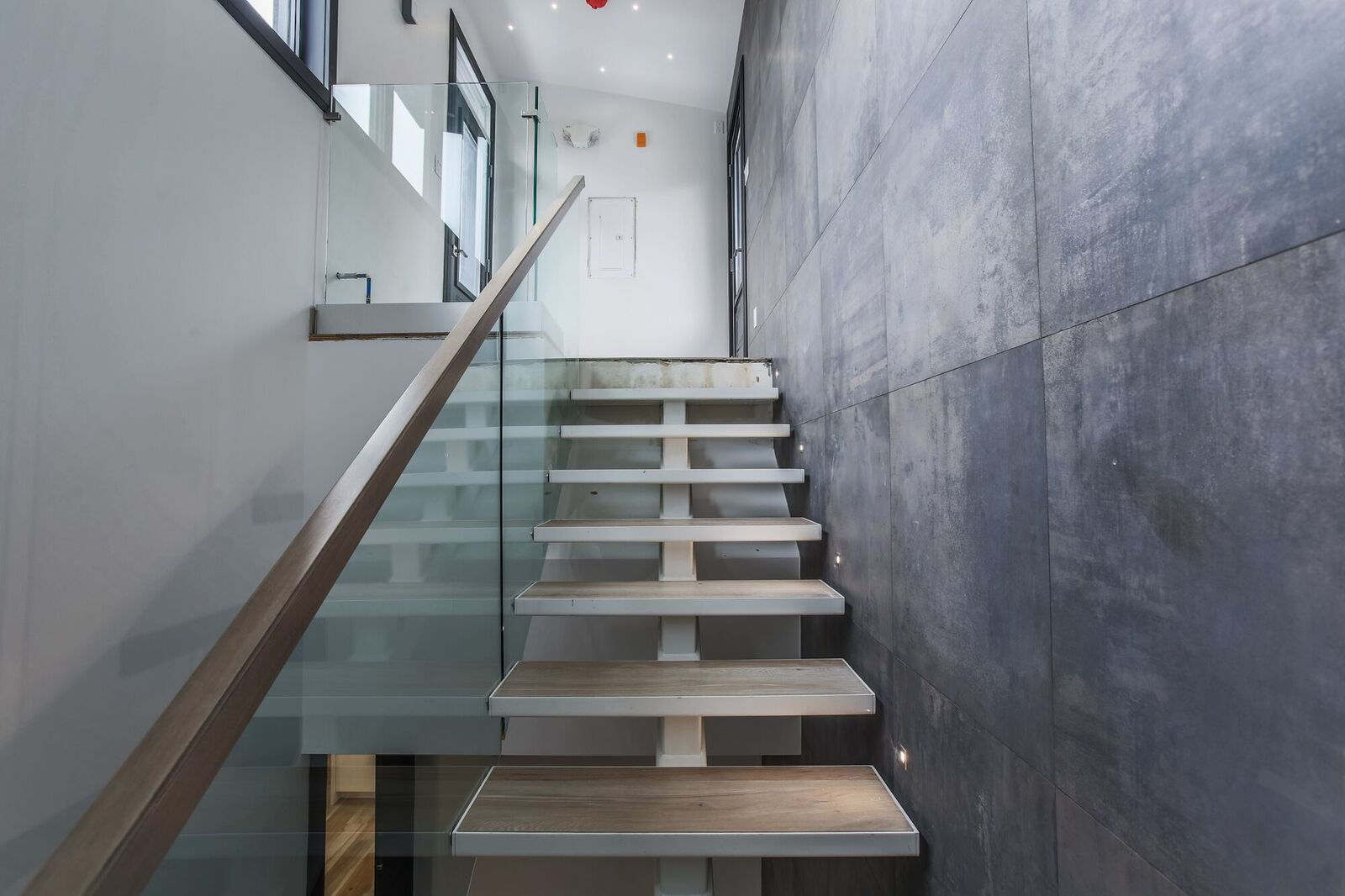 Interior view of a modern building with dark tiled floor, white walls, glass railing, and a staircase with wooden steps and white supports, leading to an upper floor with large windows and a white door.