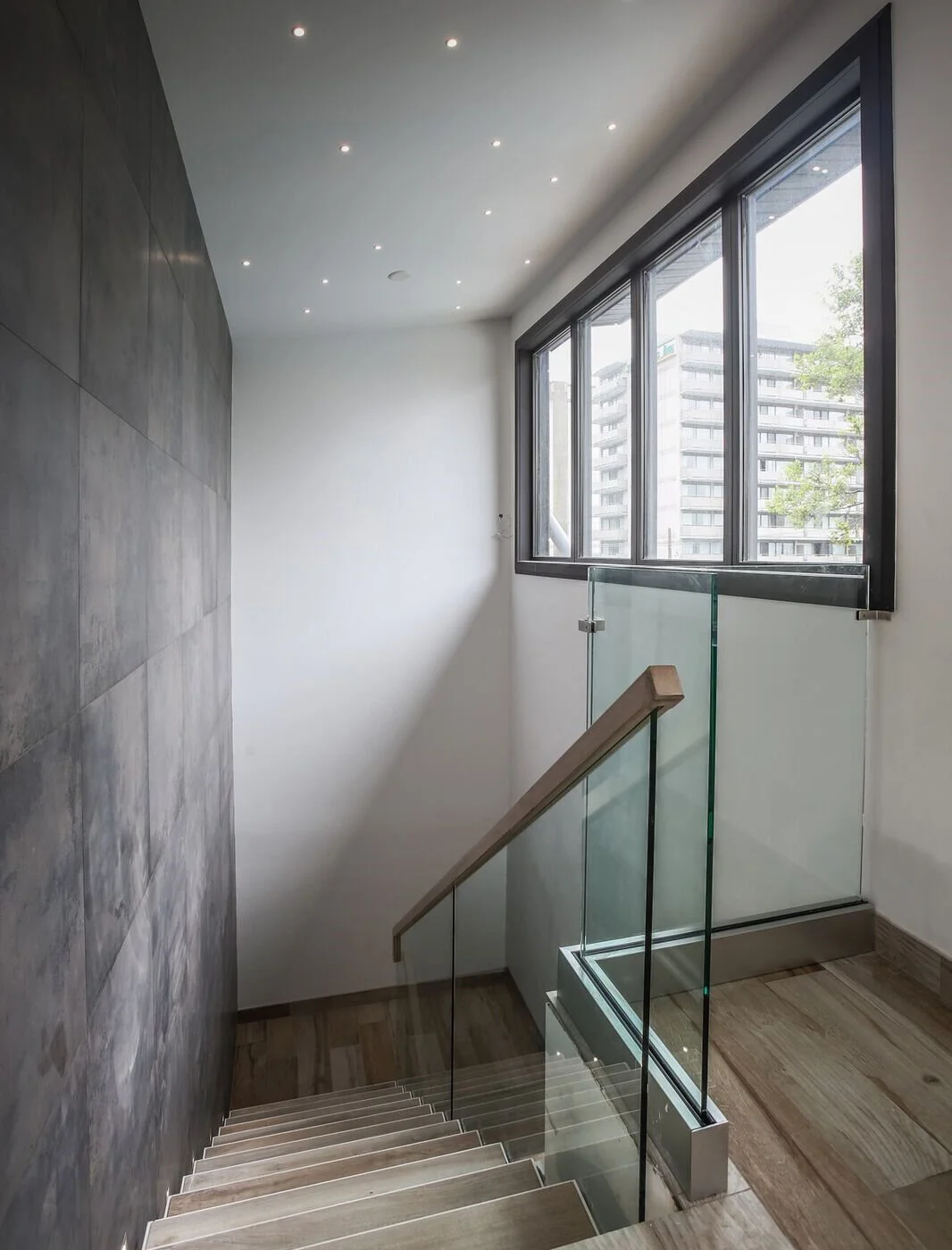 Modern interior staircase with wooden steps, glass railing, large window, and gray wall panels.