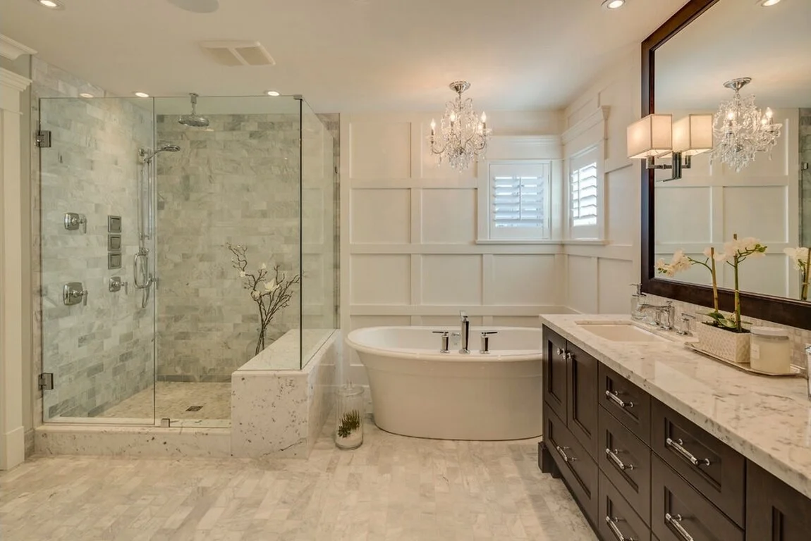A luxurious bathroom featuring a glass-enclosed shower with marble tiles, a standalone bathtub, a dark wood vanity with a marble countertop, and elegant chandeliers and wall-mounted lights.