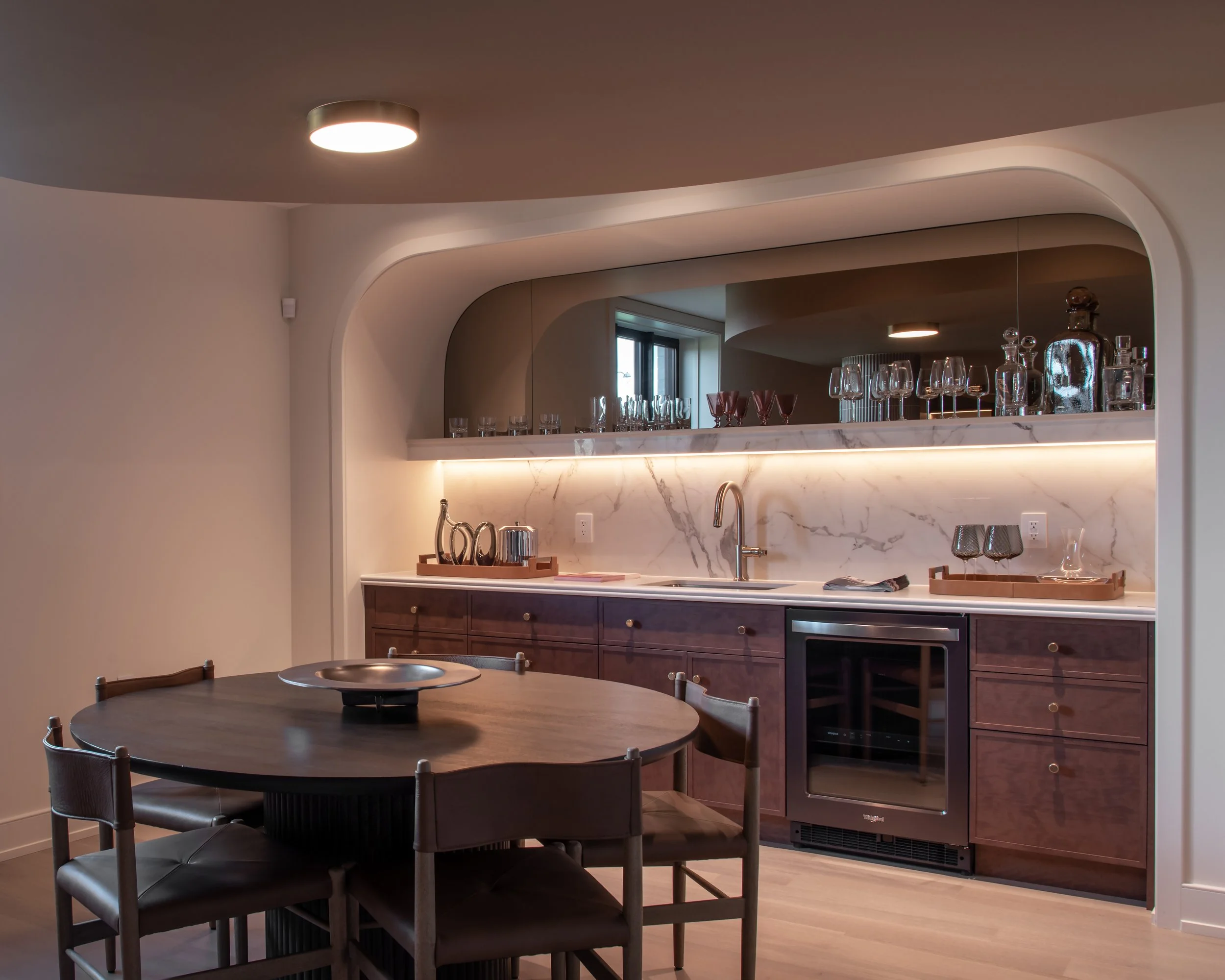 Modern kitchen with wooden cabinets, marble backsplash, round dining table with six chairs, under-cabinet lighting, glassware on a shelf, stainless steel wine cooler, and a mirror reflecting a window.