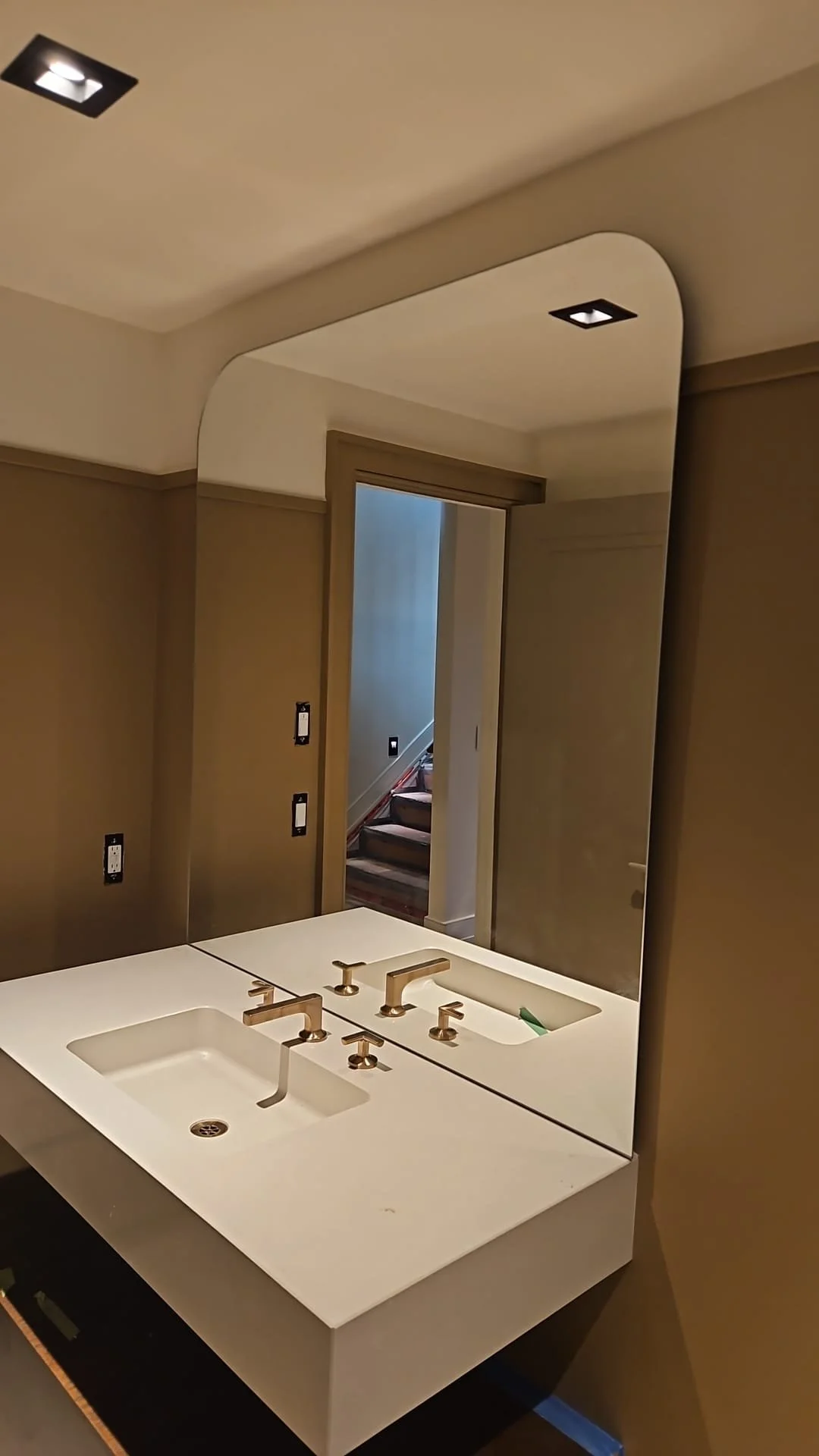 Bathroom vanity with a white countertop, dual sinks, and gold faucets, reflected in a large mirror, with a doorway leading to stairs in the background.