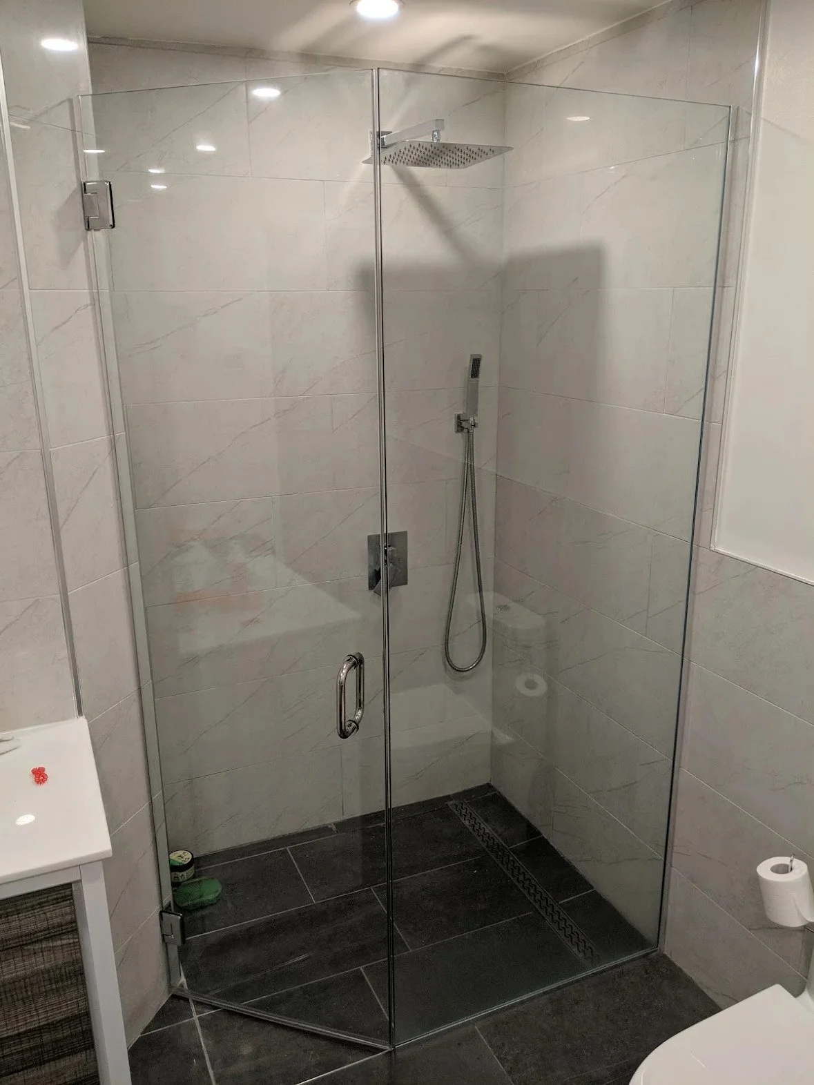 Modern bathroom with a clear glass shower enclosure containing a rainfall showerhead and handheld shower. The walls are tiled with large, light-colored tiles, and the floor has dark tiles with a linear drain. A toilet is partially visible in the corner, and a small white cabinet is next to the shower.