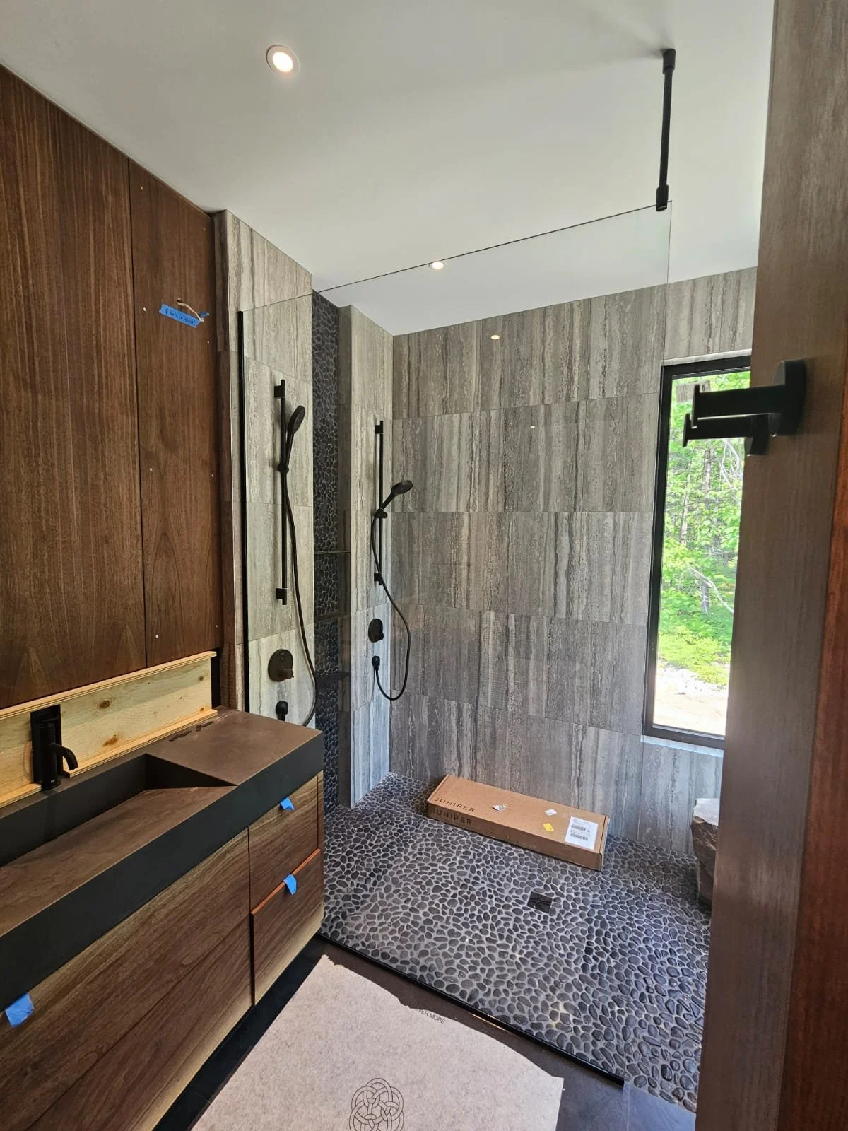 Bathroom shower area with two handheld showerheads, stone tile walls, pebble floor, a window showing greenery outside, and a cardboard box on the floor.