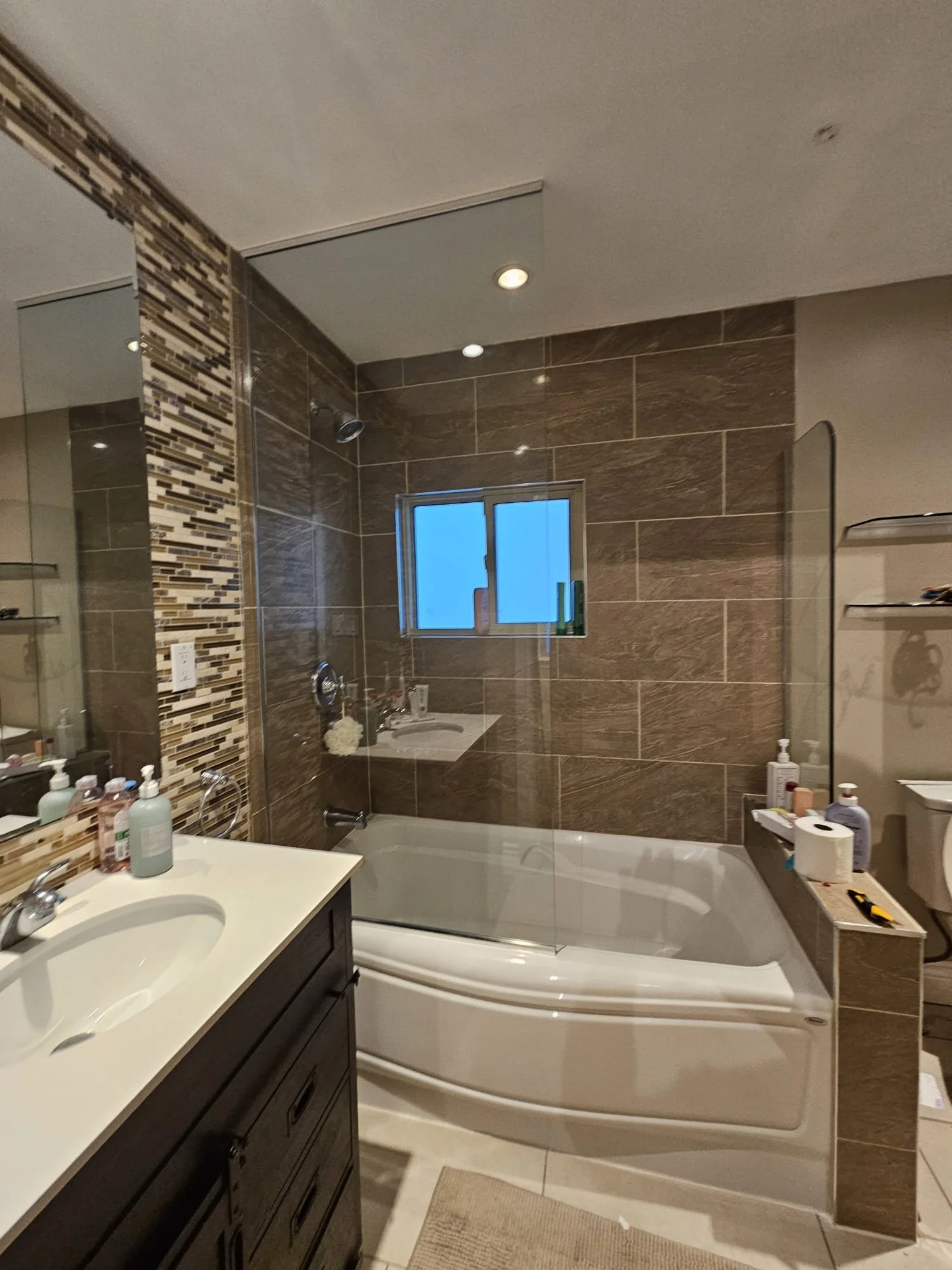 A modern bathroom with a bathtub, brown tiled walls, a small window, a white sink, and a dark vanity.