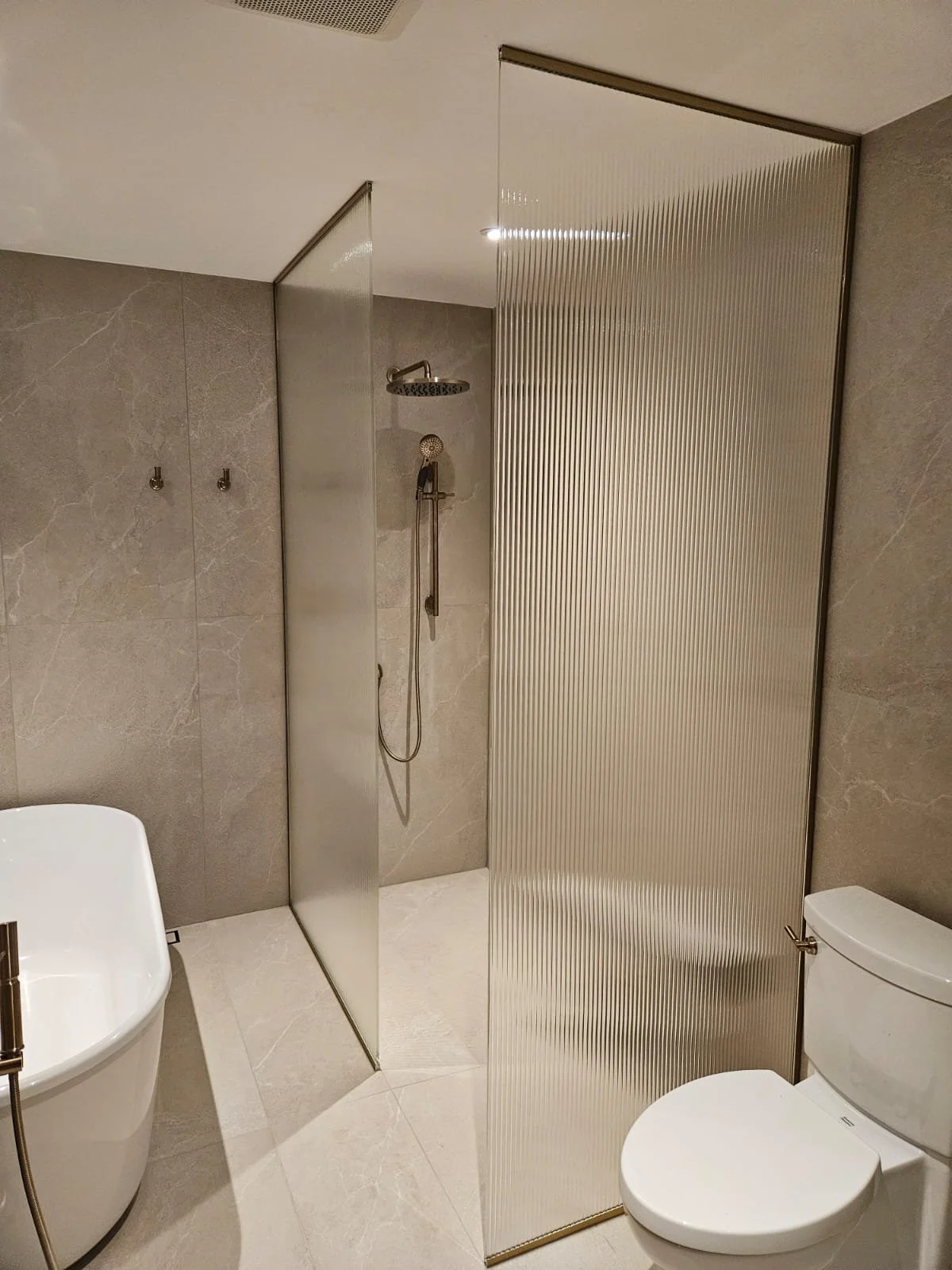 Modern bathroom with a glass shower enclosure, a white freestanding bathtub, and a white toilet. The shower has a dual partition wall, one clear and one ribbed, with a showerhead and handheld sprayer on the beige tiled wall.