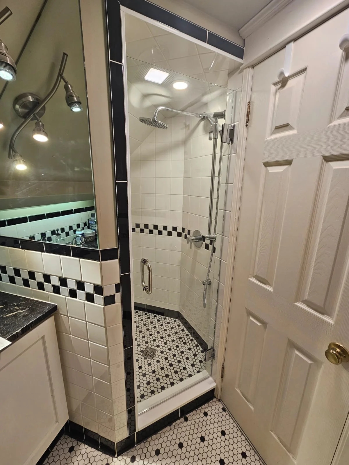 A small glass shower enclosure with black and white hexagonal tile flooring and white tile walls with a black and white checkered border; a ceiling-mounted shower head and adjustable hand-held shower; beige door with gold knob, and part of a bathroom