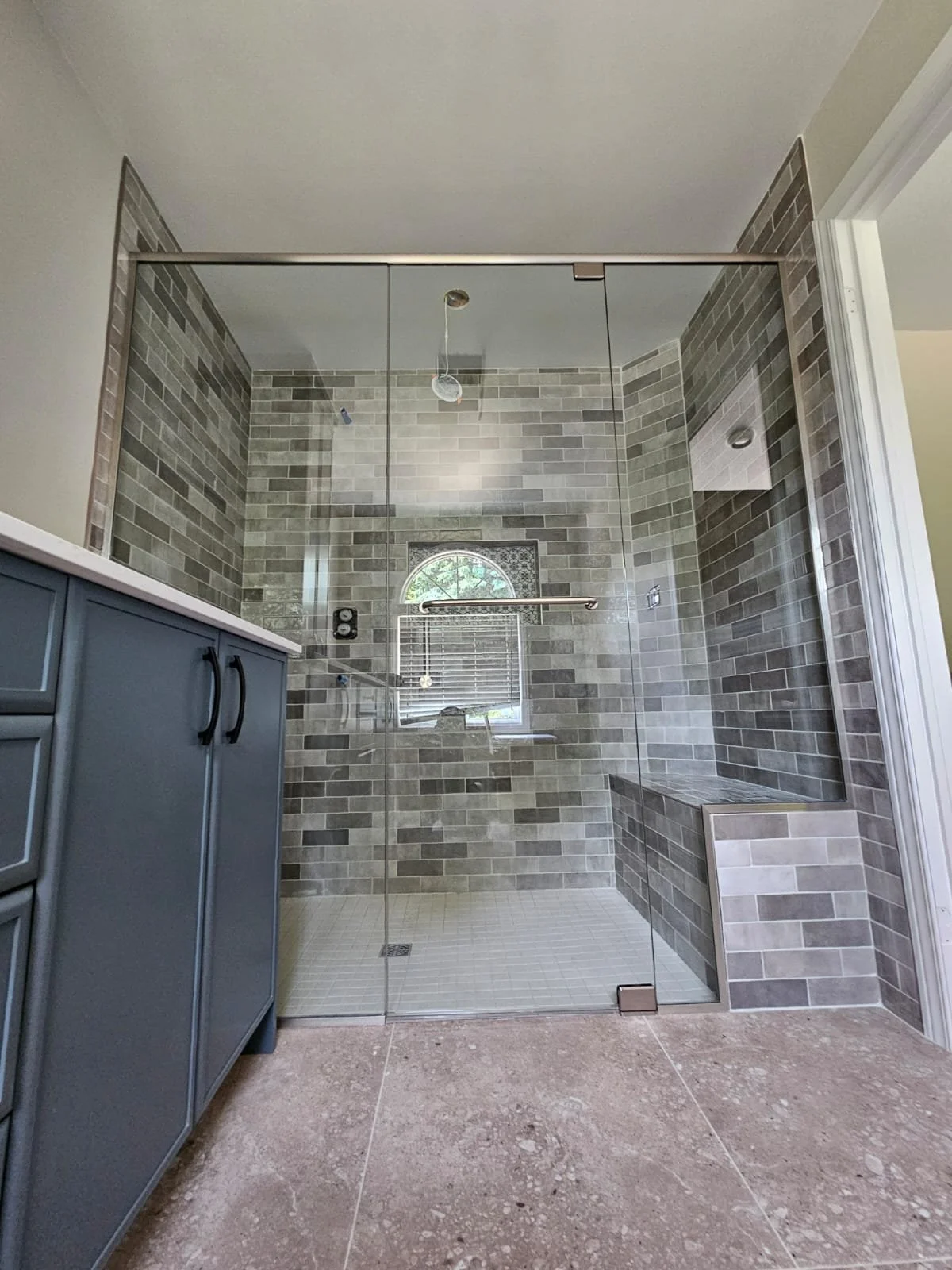 Modern bathroom with a glass shower enclosure, gray tiled walls, a window with an arched top, and a blue vanity with black handles.