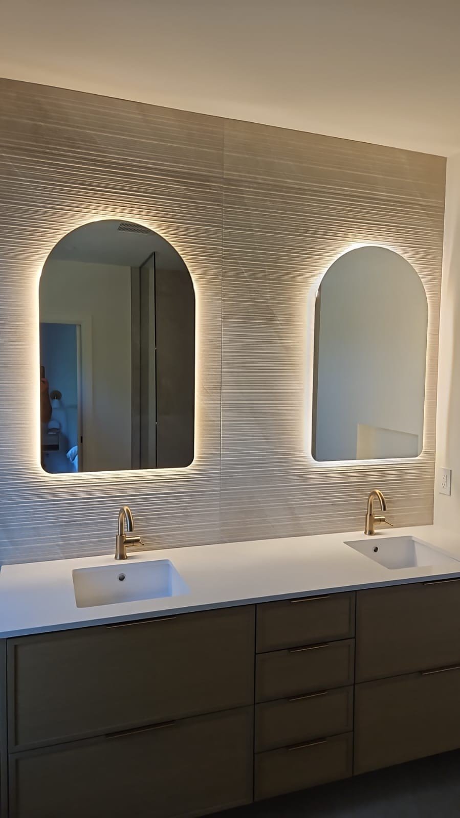 Modern bathroom vanity with two sinks, brass faucets, and two illuminated mirrors on textured wall.