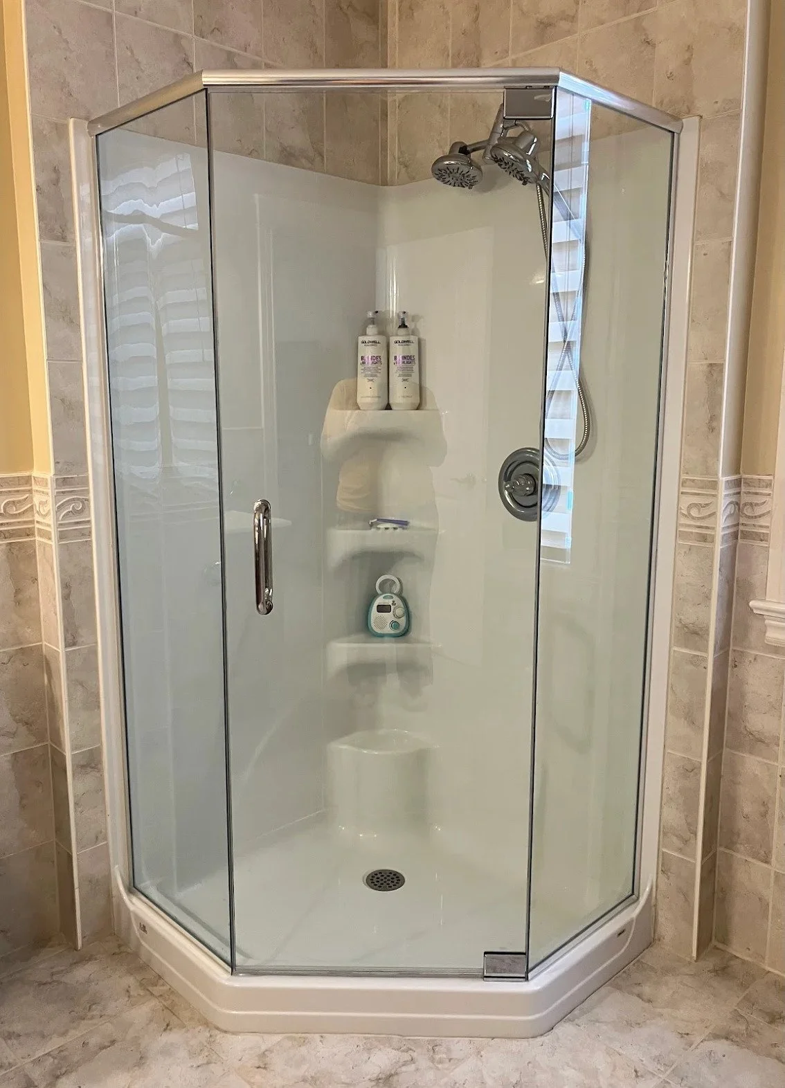 A glass-enclosed shower with two showerheads and shelves inside, containing bottles of shampoo and body wash, and a timer or speaker.
