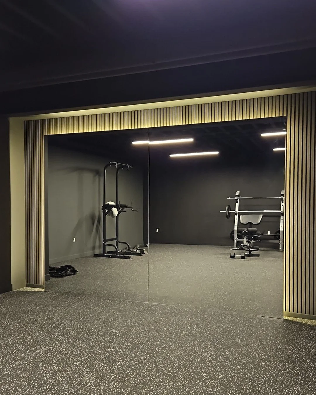 Empty gym room with black walls, rubber flooring, a weight rack, and a mirror reflecting the room.