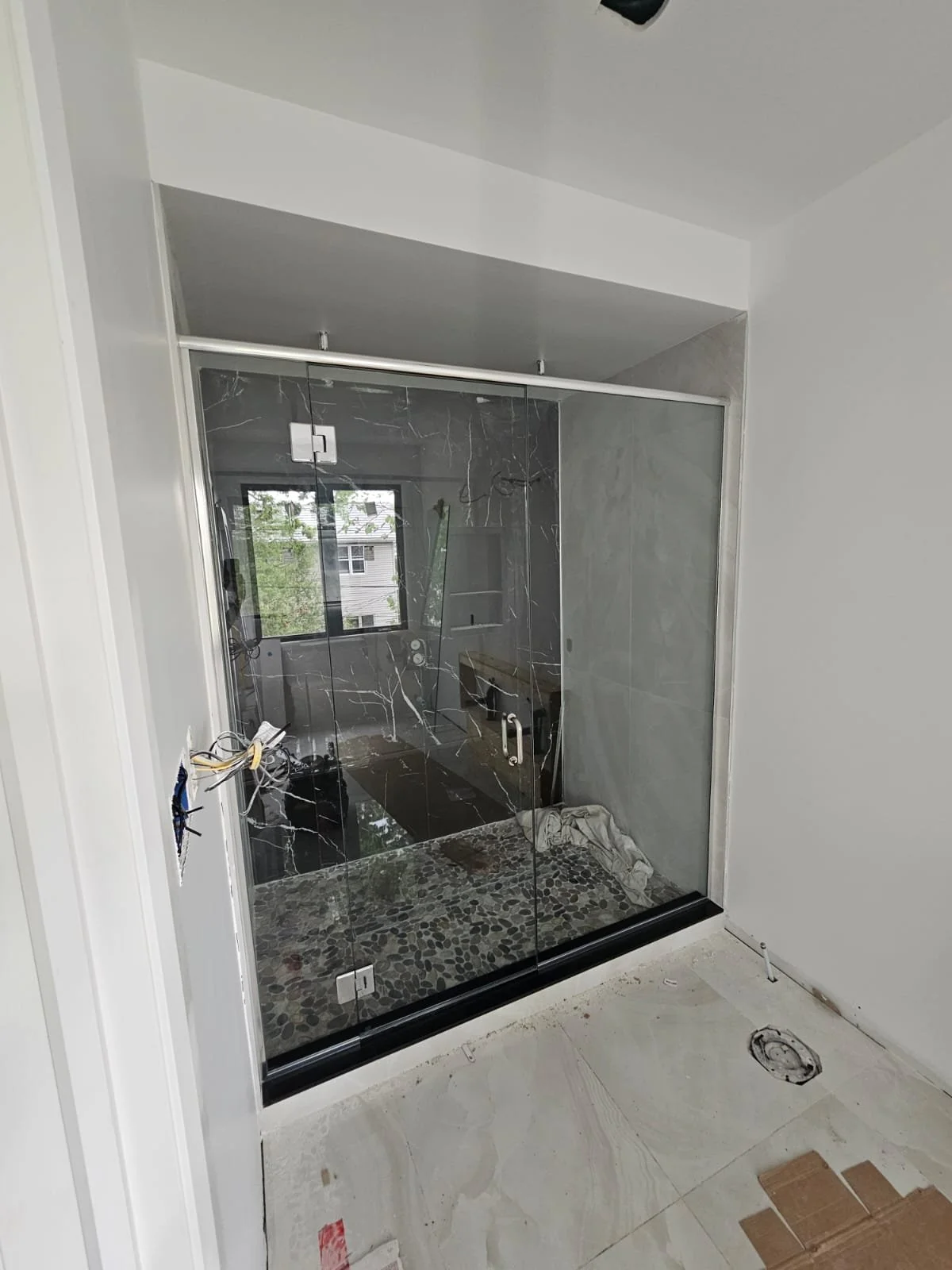 A glass shower enclosure with a black marble appearance, installed in a bathroom under construction. The outside is white, and there is an electrical outlet on the wall and some construction materials on the floor.