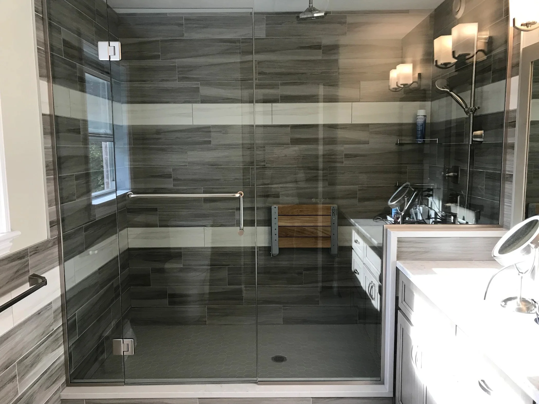 Modern bathroom with a glass shower enclosure, dark gray tiles, a window, a towel bar, and bathroom vanity with a sink and mirror.