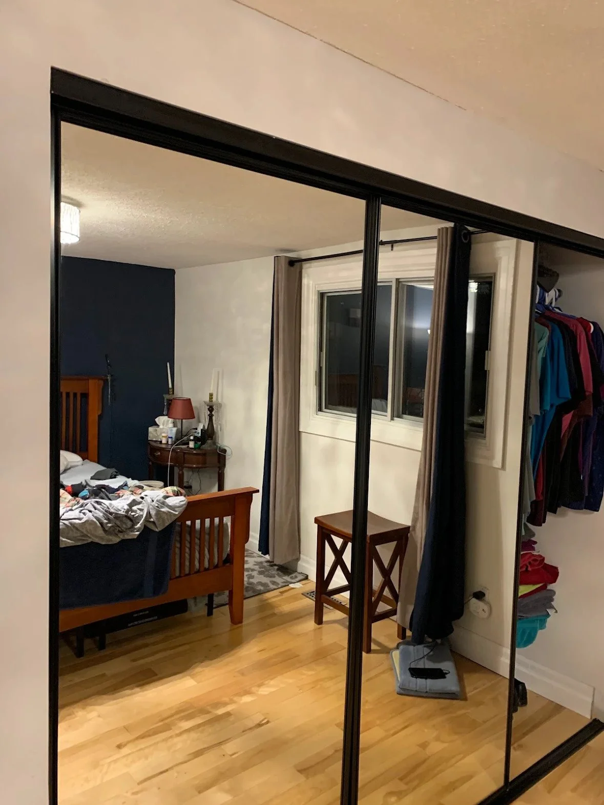 View of a bedroom with a wooden bed, bedside table with a lamp, window with curtains, and open closet with clothes.