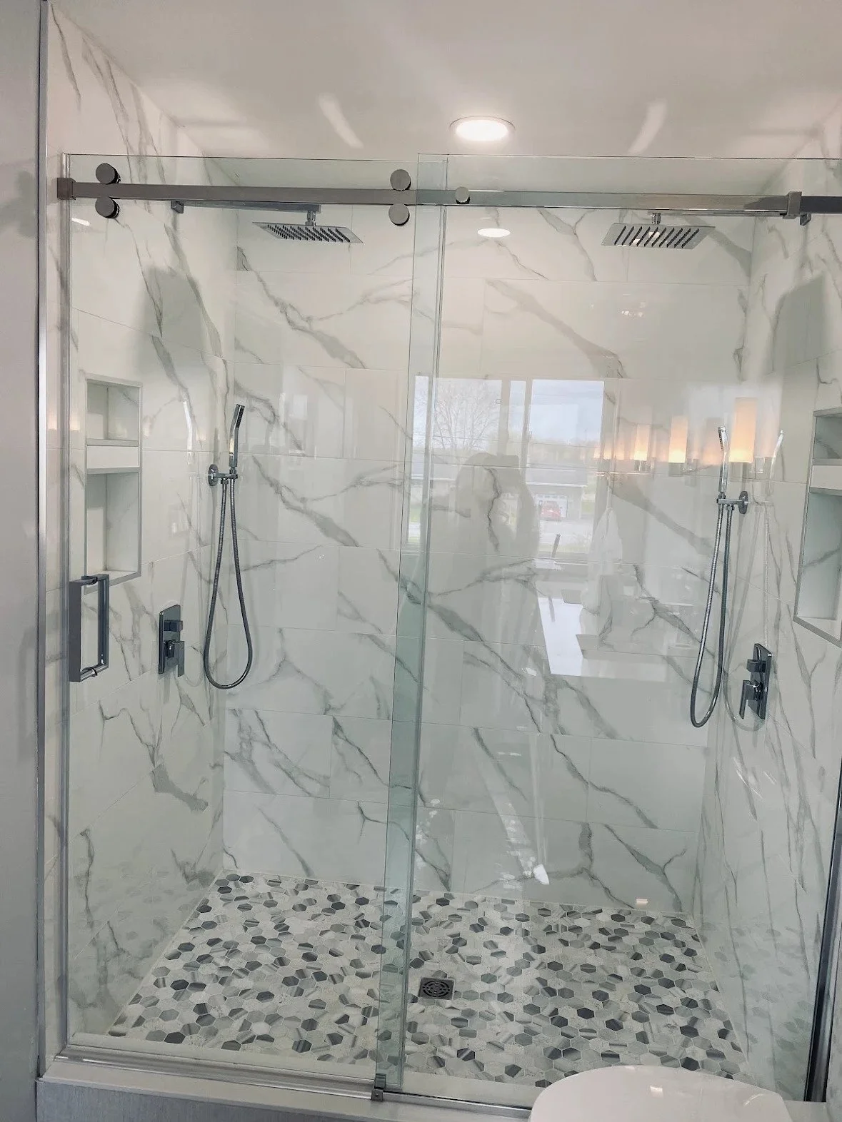 A modern walk-in shower with glass doors, marble tiles, and pebble stone flooring, featuring two shower heads and built-in shelves.